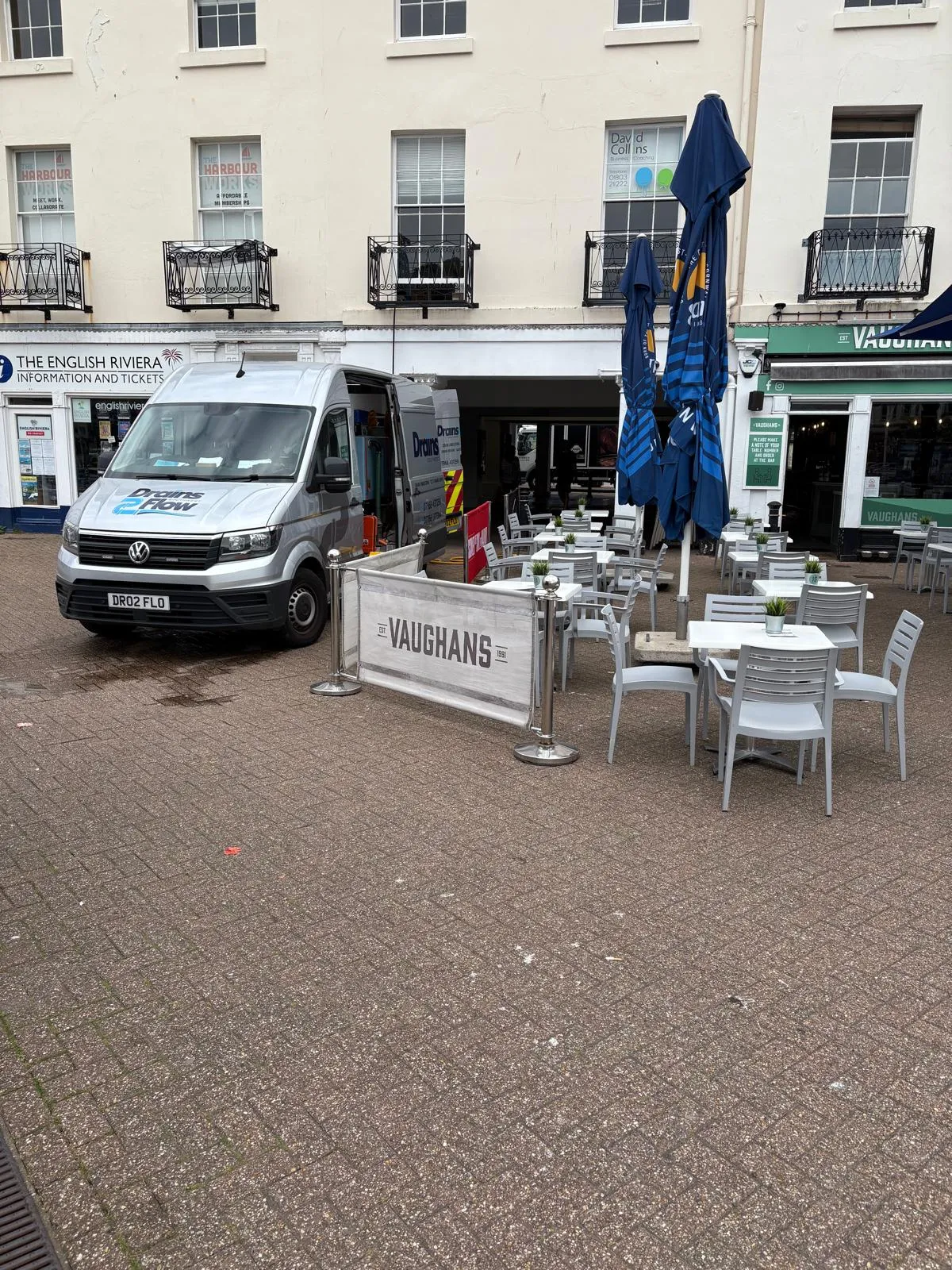 A silver Drains2Flow van with its side door open, parked on a paved area outside shops and a restaurant in Crediton, Devon.