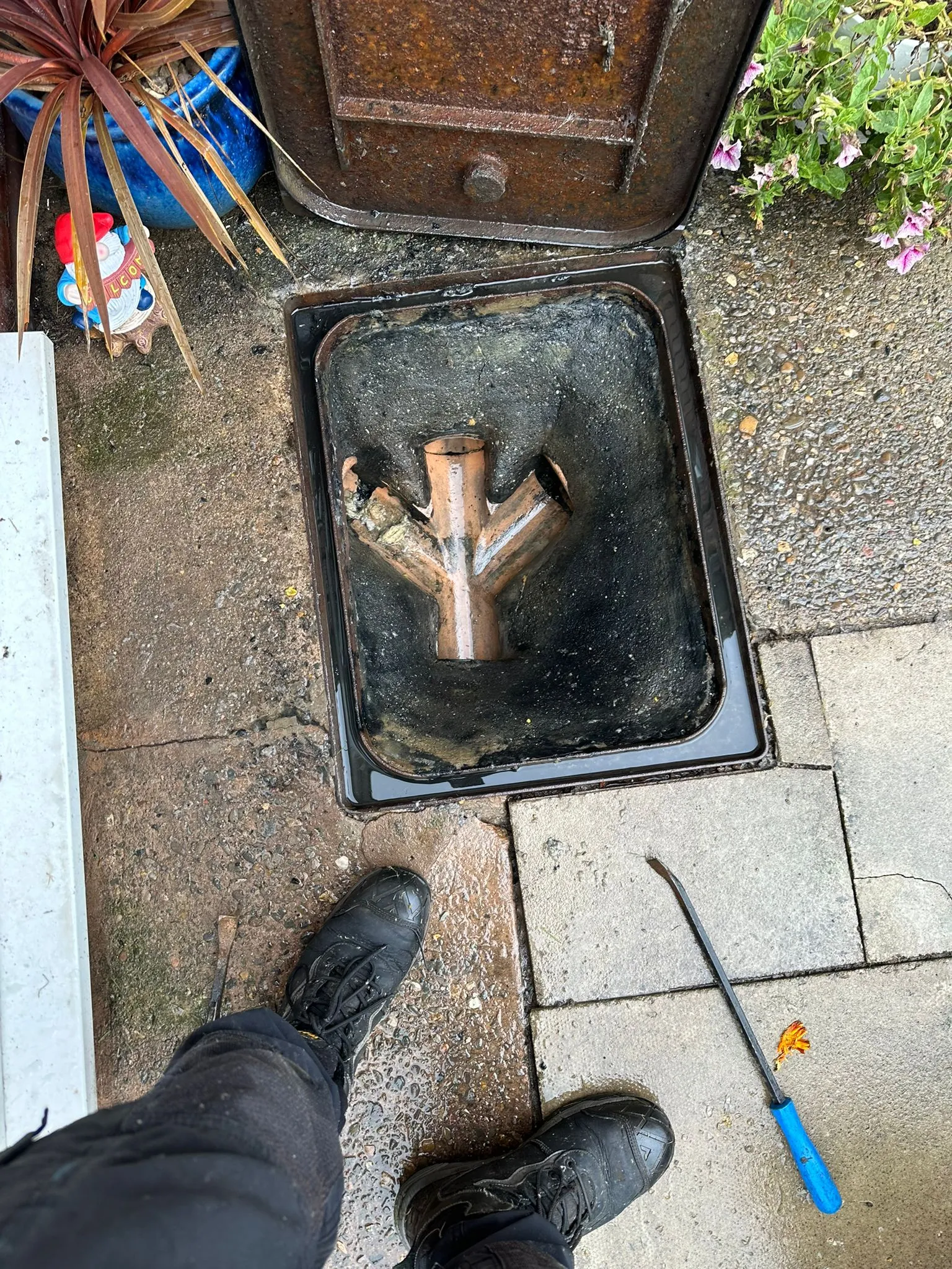 Drainage engineer's feet and tools next to an open outdoor manhole during a drain unblocking service.