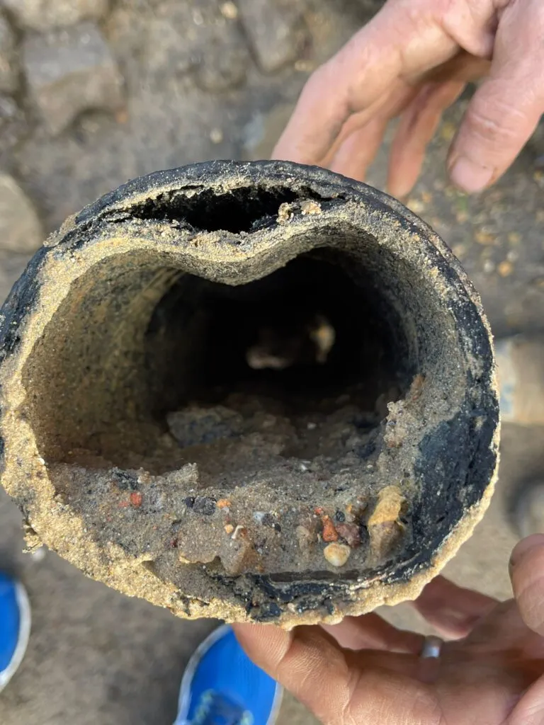 Hands holding a cross-section of a badly damaged and blocked pitch fibre drain pipe, revealing severe internal narrowing due to debris and structural issues.