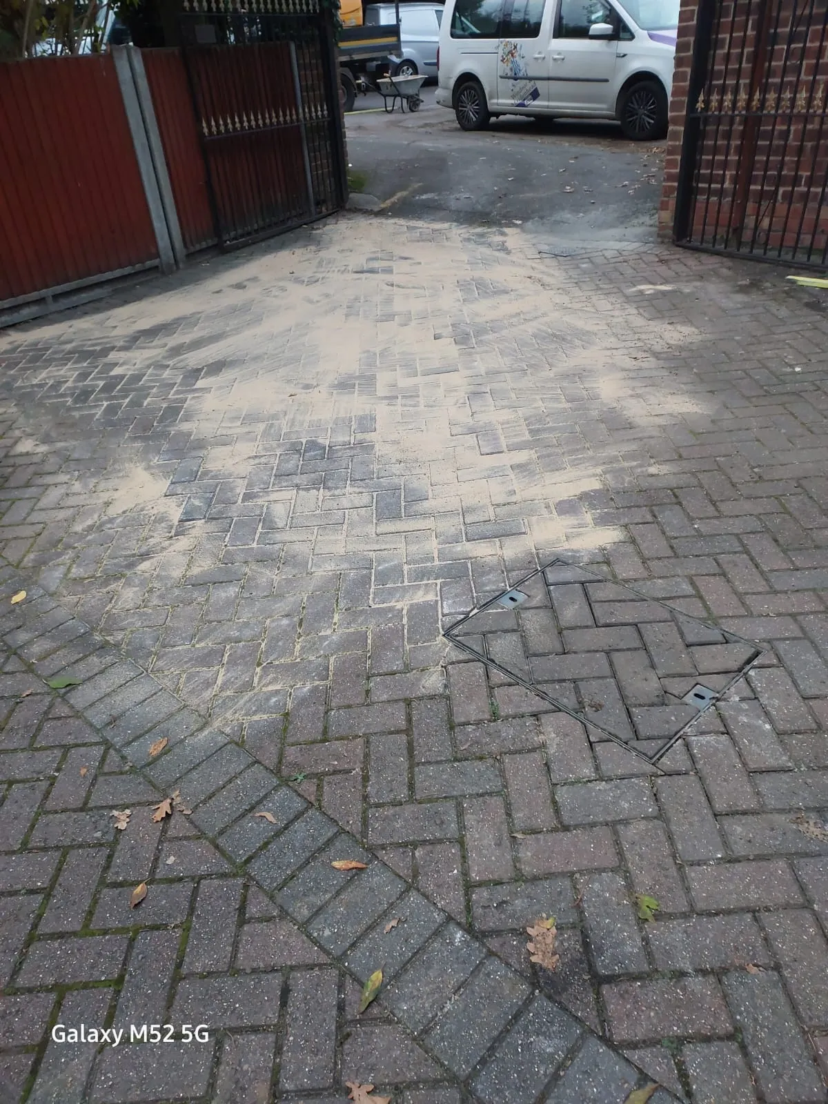 A paved driveway with sand and an open drain cover frame, suggesting recent drainage system repairs or installation.
