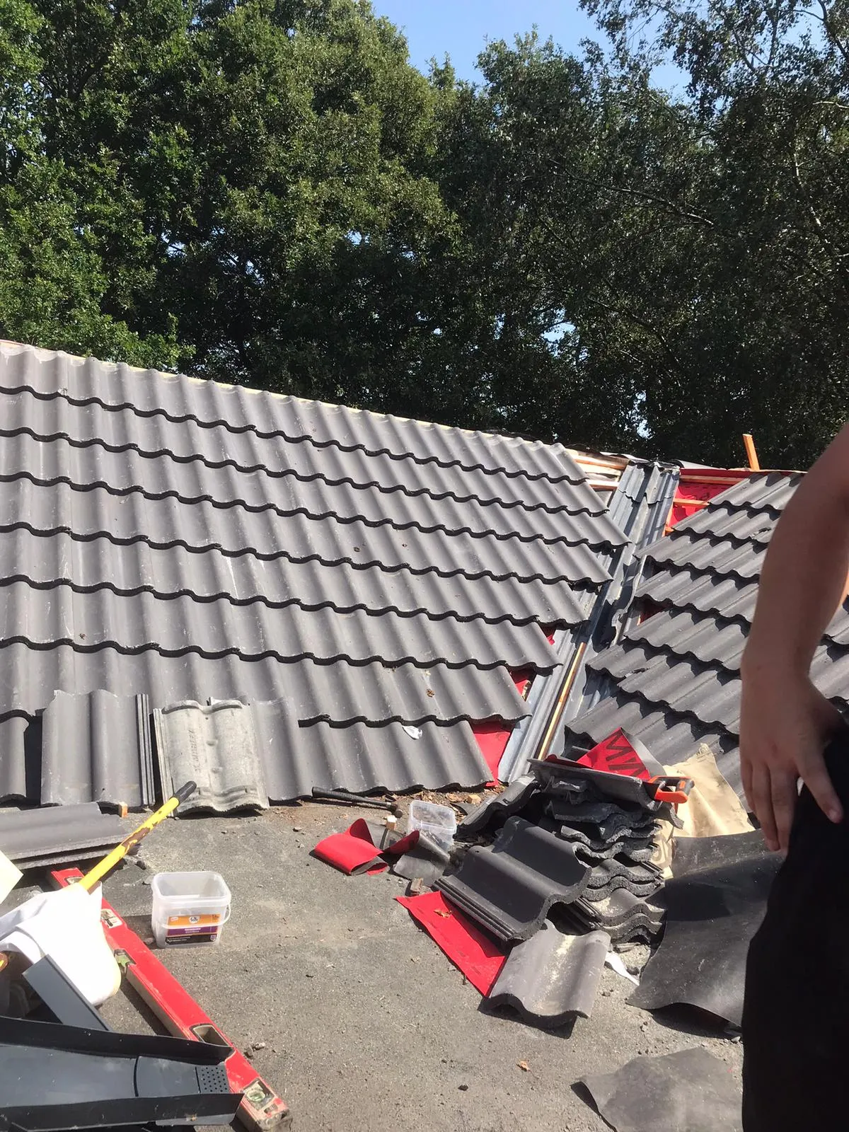 A partial view of a residential roof undergoing renovation, with new grey interlocking tiles in place, red underlayment visible, and construction debris and tools on the roof.
