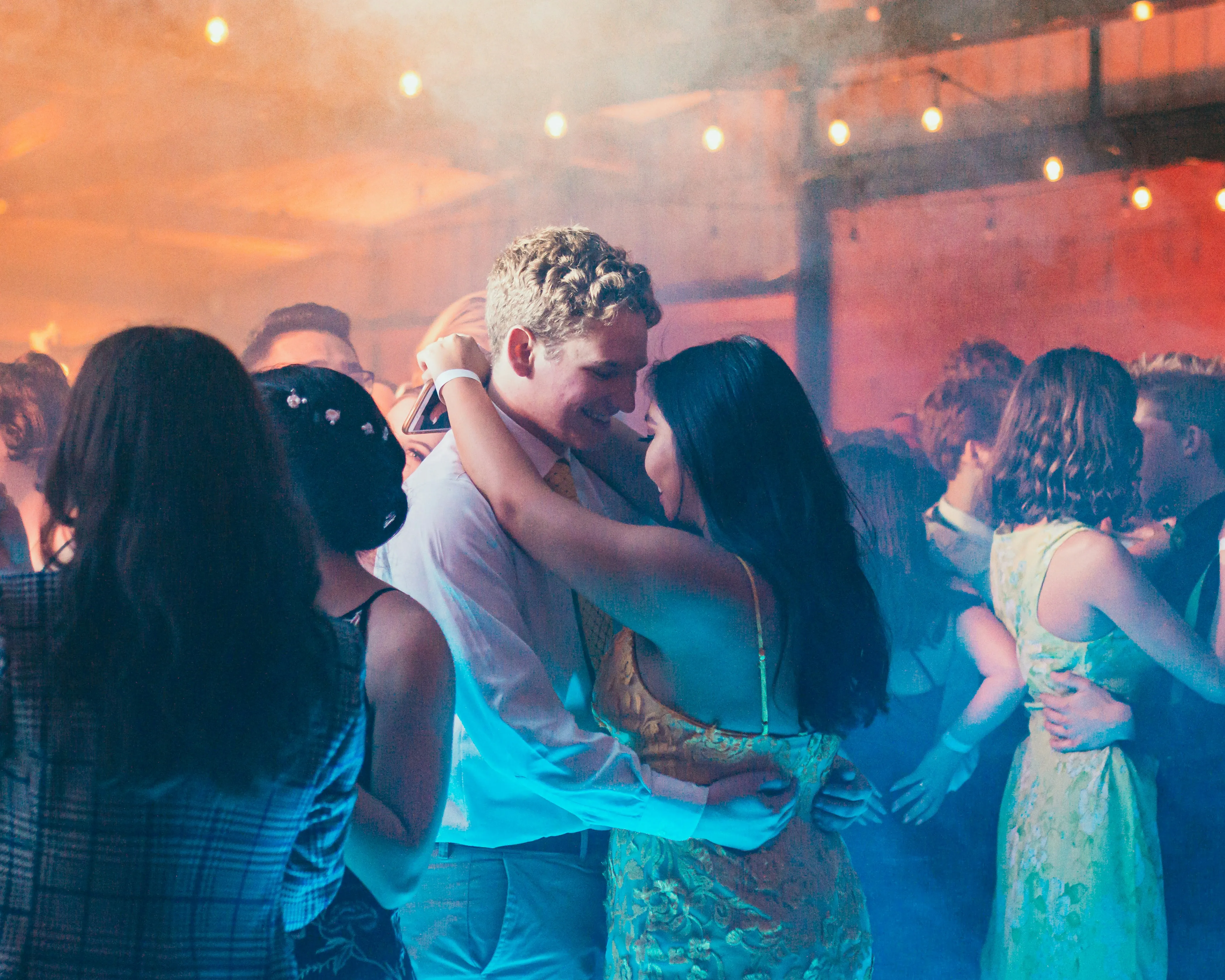 A couple slow dancing on a party dance floor, bathed in warm and cool lighting with other guests in the background, creating a joyful, unforgettable vibe.