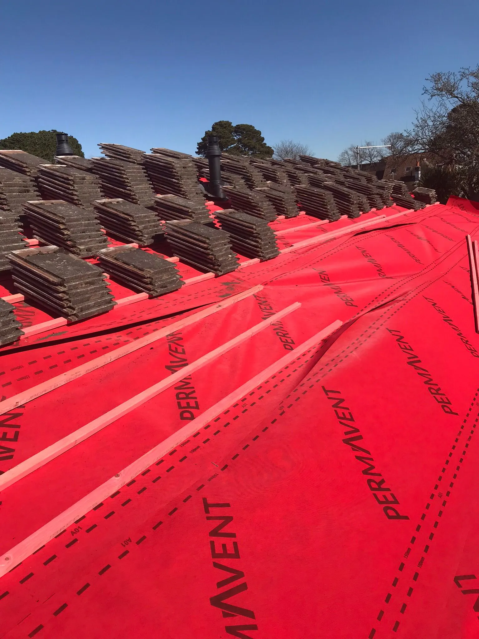 Pitched roof installation in progress, showing red breathable membrane, wooden battens, and stacks of new roof tiles.