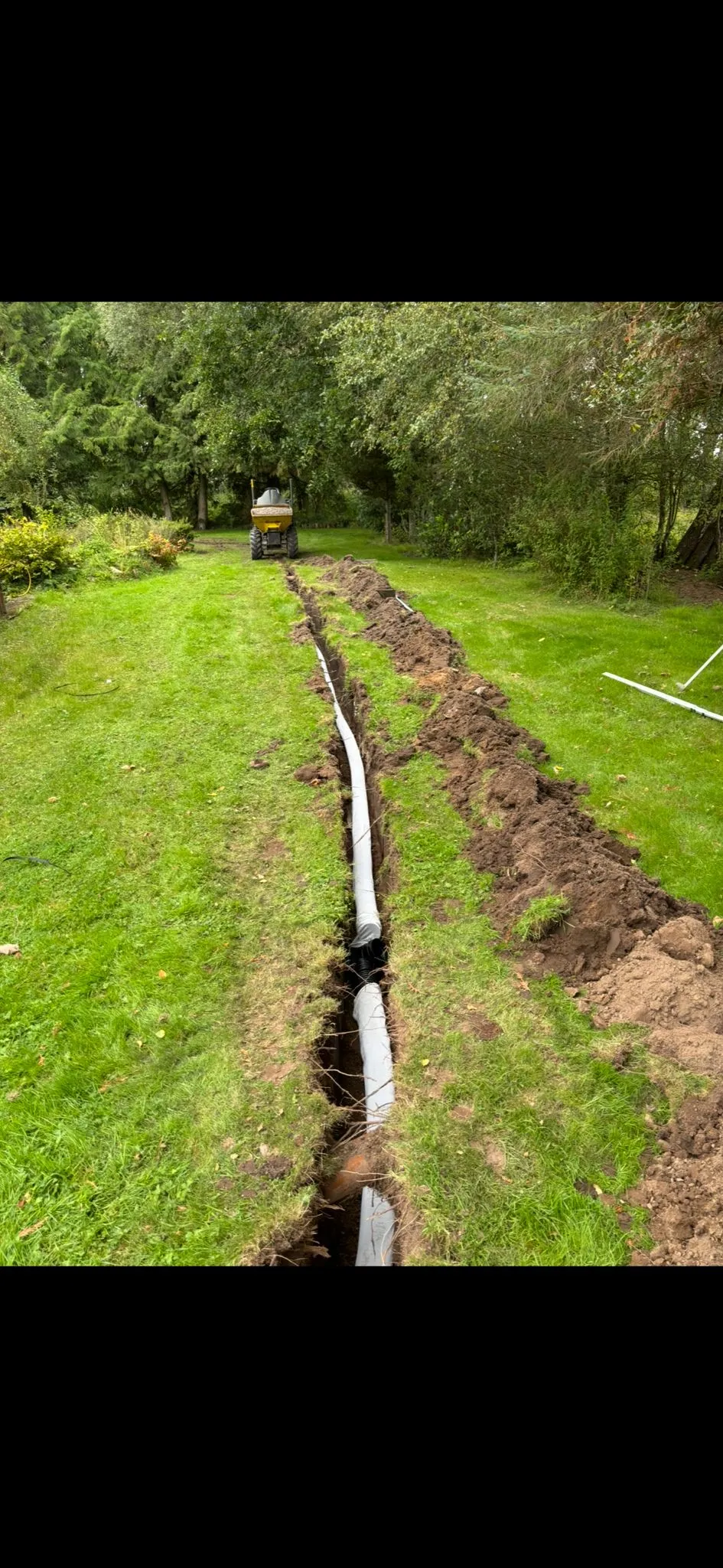 An excavated trench with new drainage pipes being laid in a garden, with a dumper in the background.