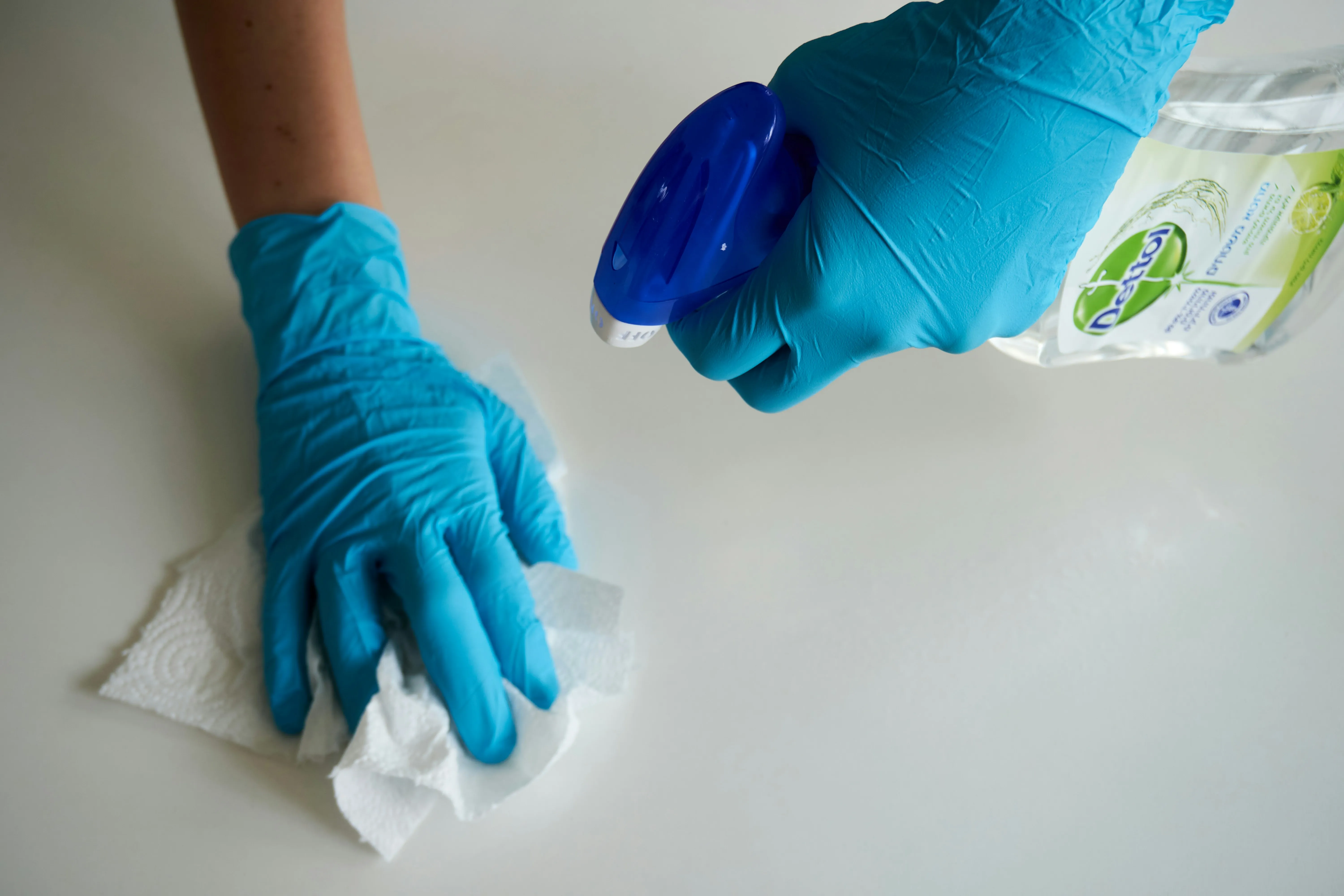 A person wearing blue gloves cleaning a white surface with a disinfectant spray bottle and a paper towel.