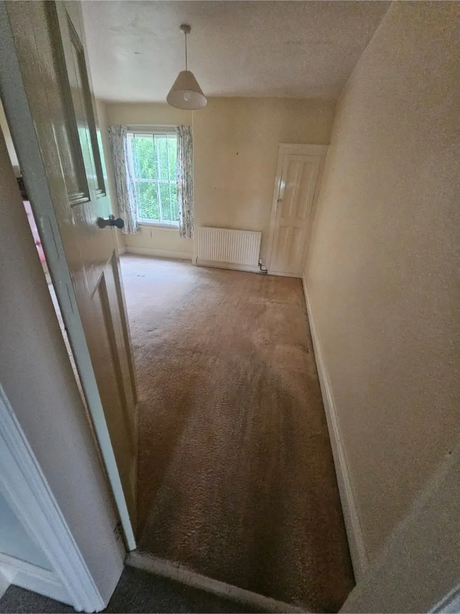 Empty bedroom with brown carpet, window, and radiator after a house clearance, ready for new occupants.