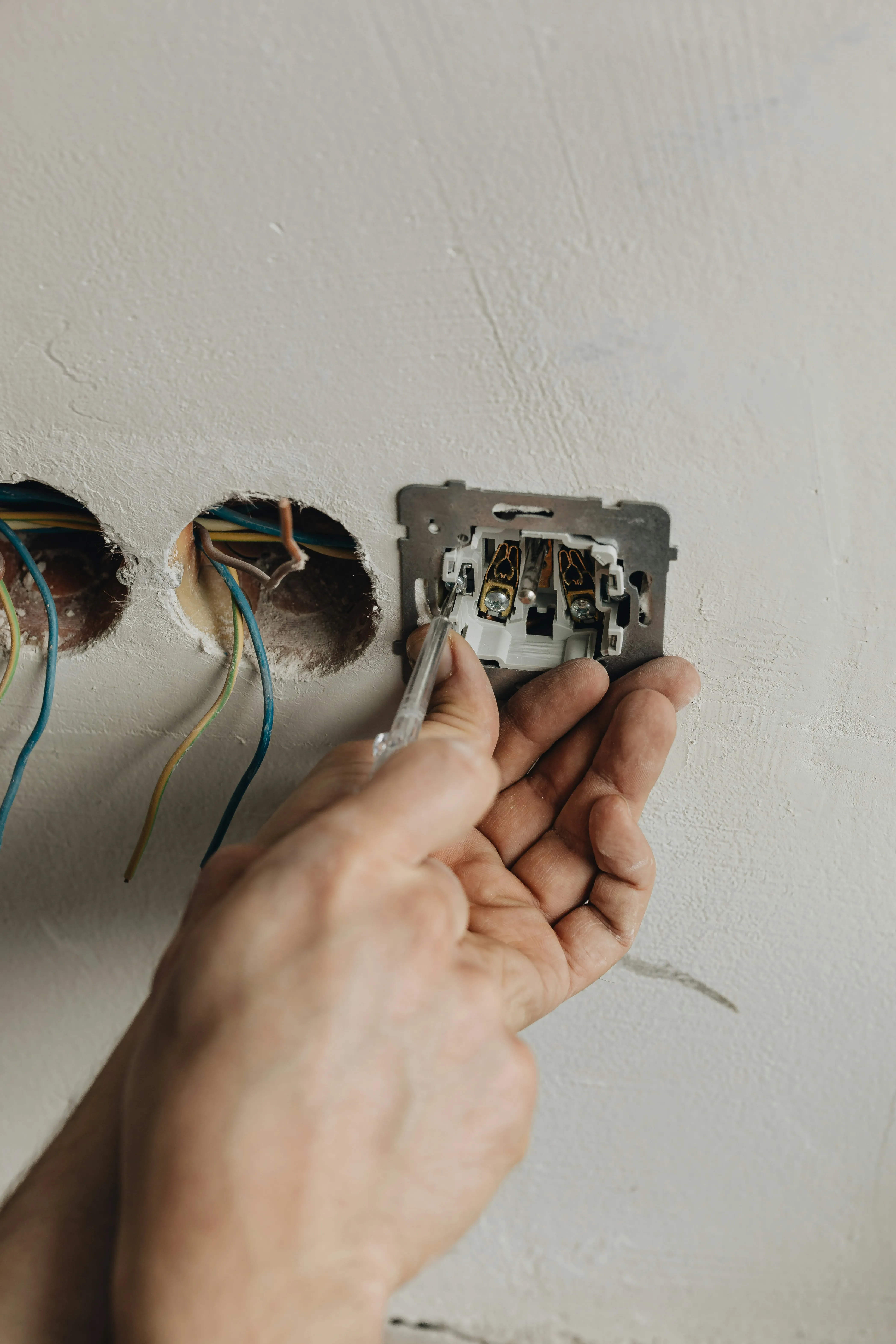 Close-up of an electrician's hands using a screwdriver to connect electrical wires to a wall socket or switch plate.