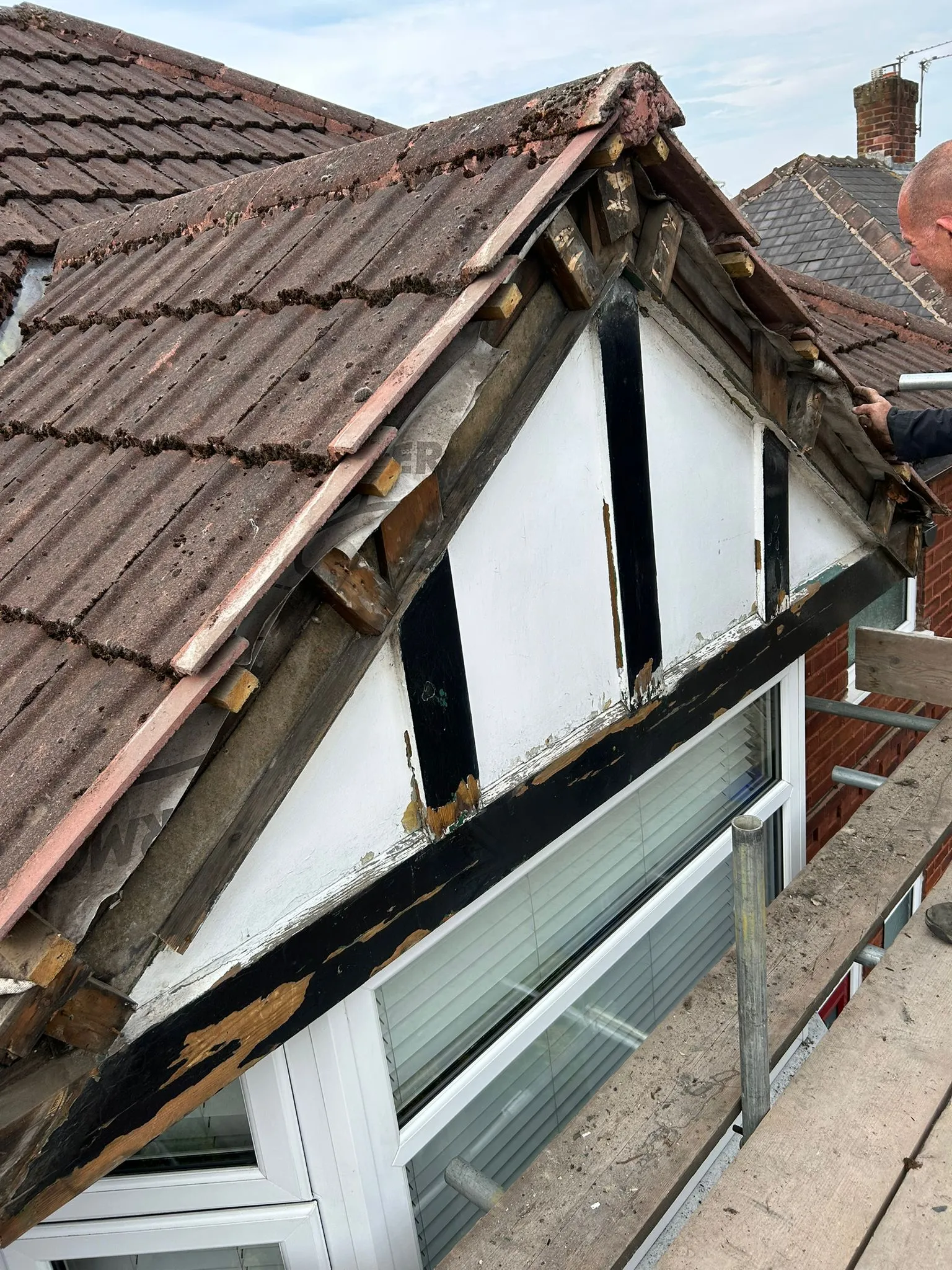 Damaged pitched roof dormer window with old tiles and exposed timber, undergoing repair by a roofer on scaffolding.