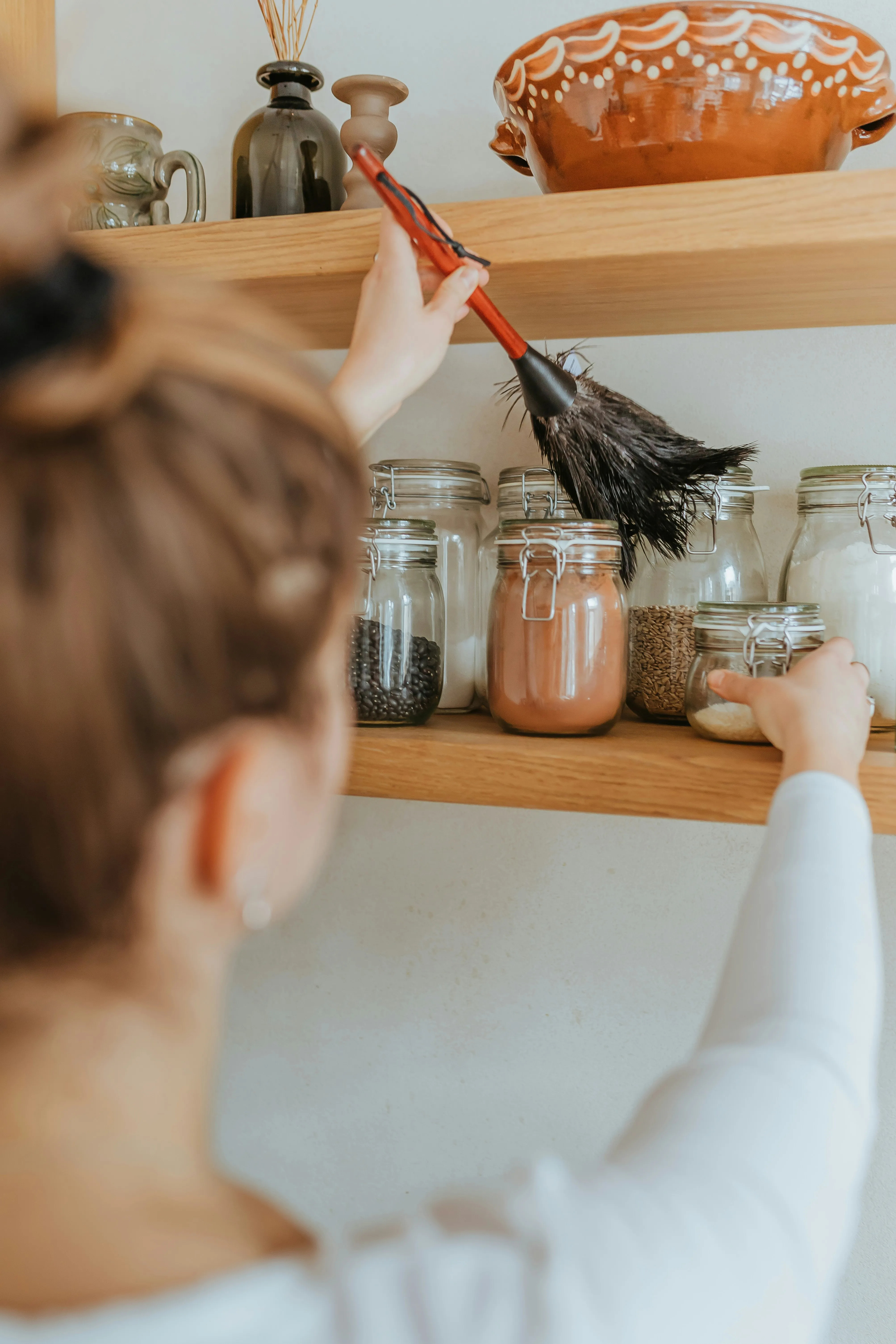 A person's arm reaching up with a feather duster to clean a wooden shelf, with several glass jars of food and decorative pottery visible.