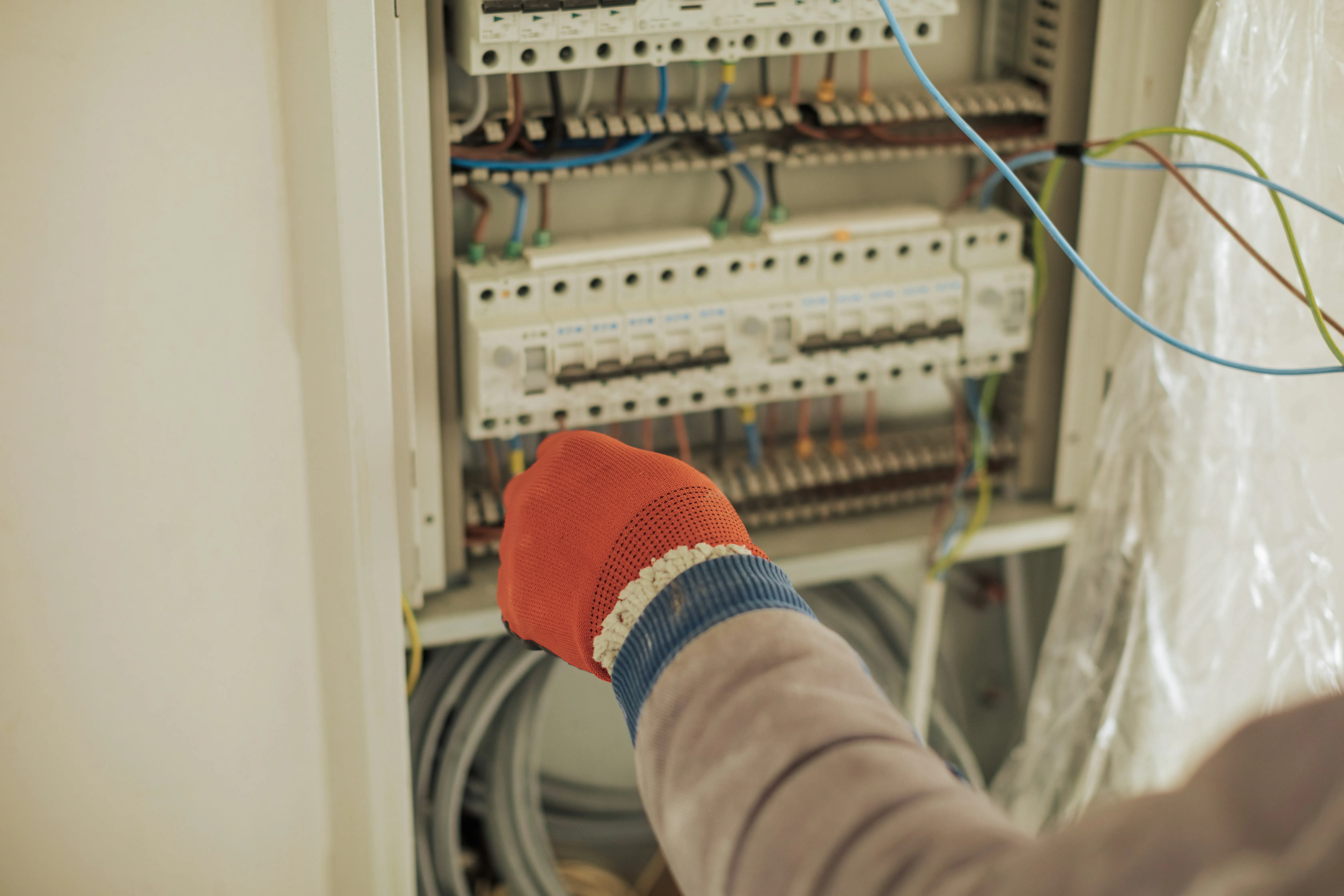 Close-up of an electrician's hand in an orange glove adjusting wires within a new consumer unit, showing circuit breakers and various coloured electrical cables.