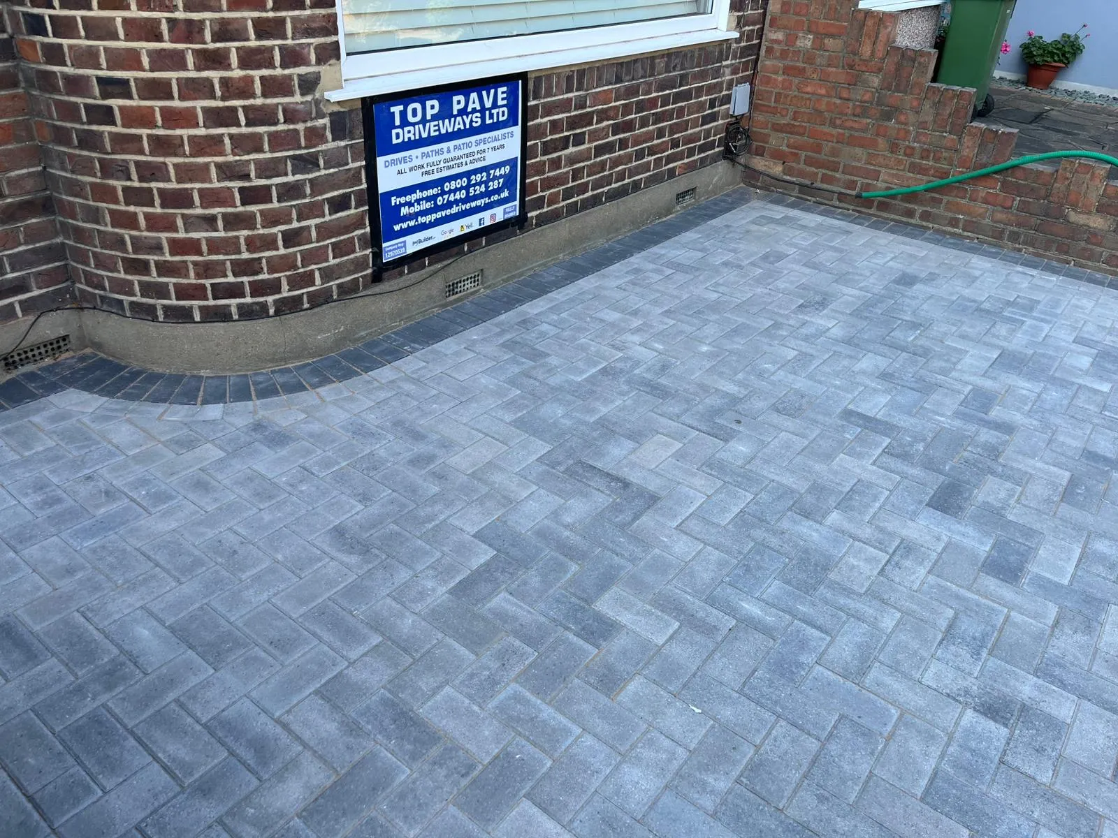 A modern grey block paving driveway or patio installed by Top Pave Driveways LTD, featuring a herringbone pattern and a contrasting dark border alongside a brick house.