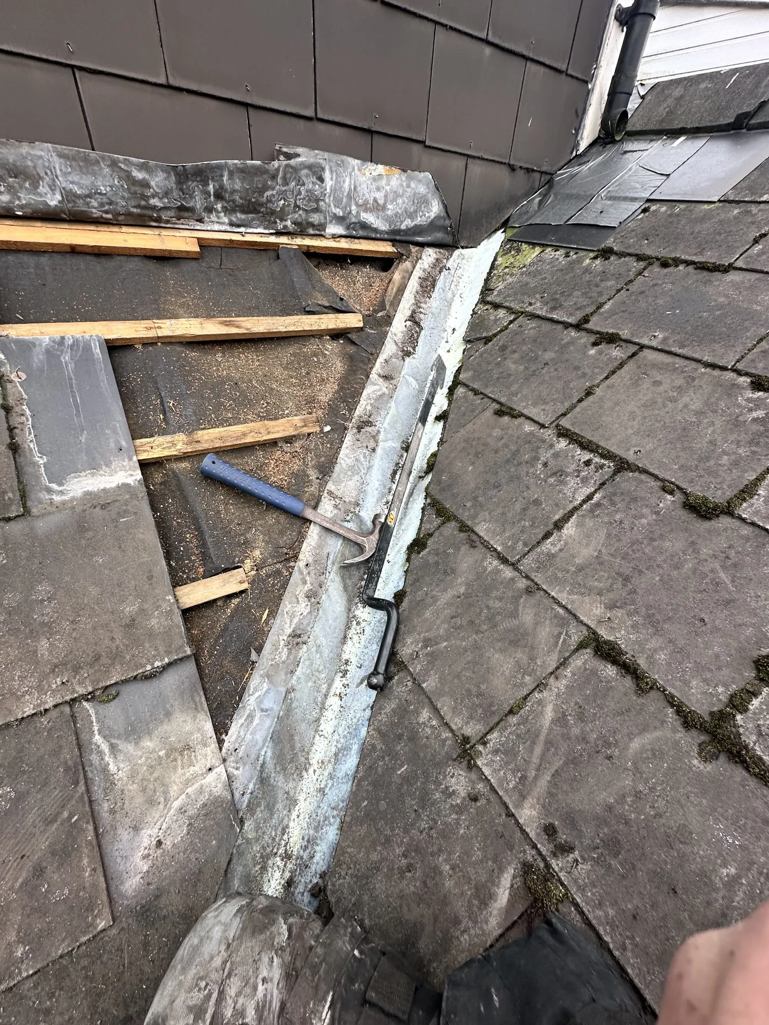 Roof repair in progress showing old mossy slates, a lead valley, exposed battens, and new slates, with roofing tools visible.