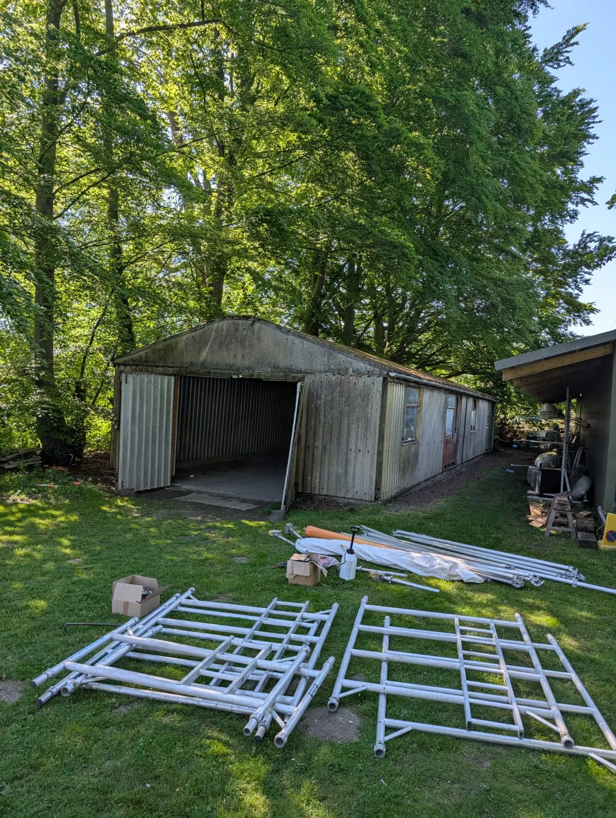 An old, long garage or shed with a grey corrugated roof and walls, with scaffolding on the grass in front, under a bright sky with green trees.