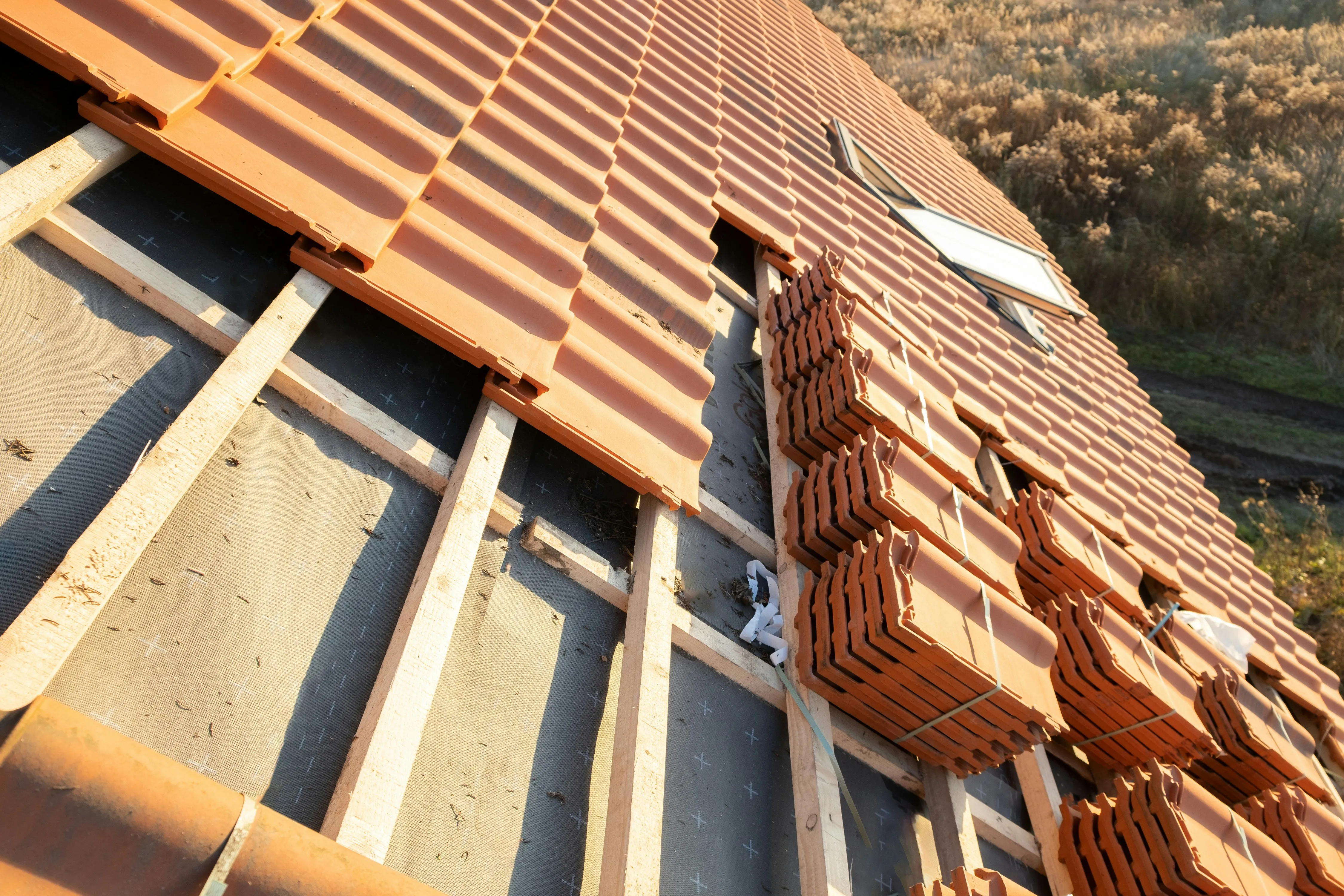 Close-up of a new pitched roof installation with terracotta tiles being laid over a breathable roofing membrane and wooden battens, with a skylight visible.