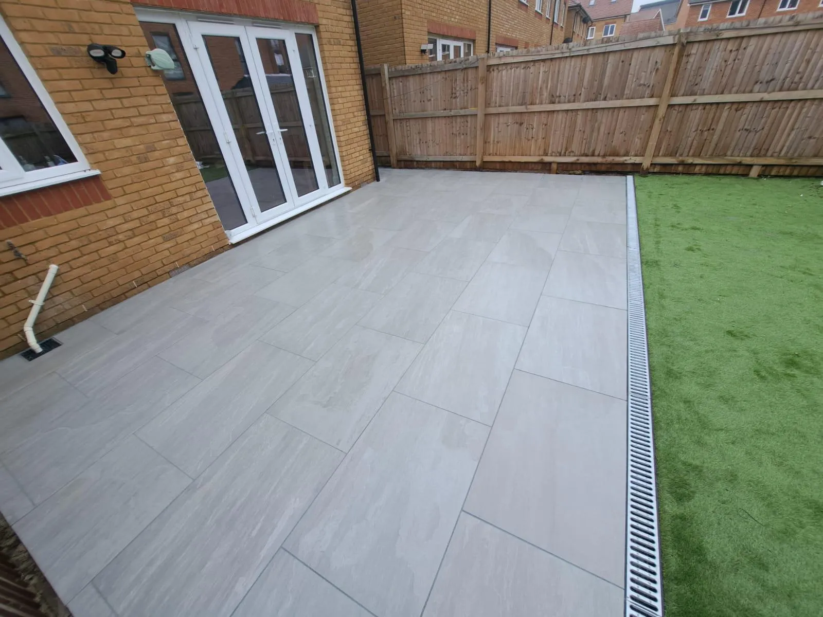 A modern grey slab patio and artificial grass in a landscaped garden, with a drainage system
