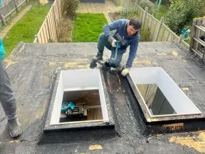 Roofer applying sealant around two skylights on a flat roof during an installation or repair project.