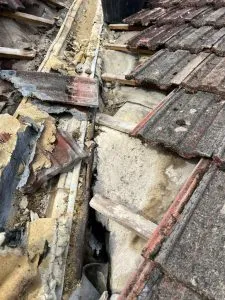 Severely damaged pitched roof with broken tiles and exposed timber structure, indicating urgent repair is needed.