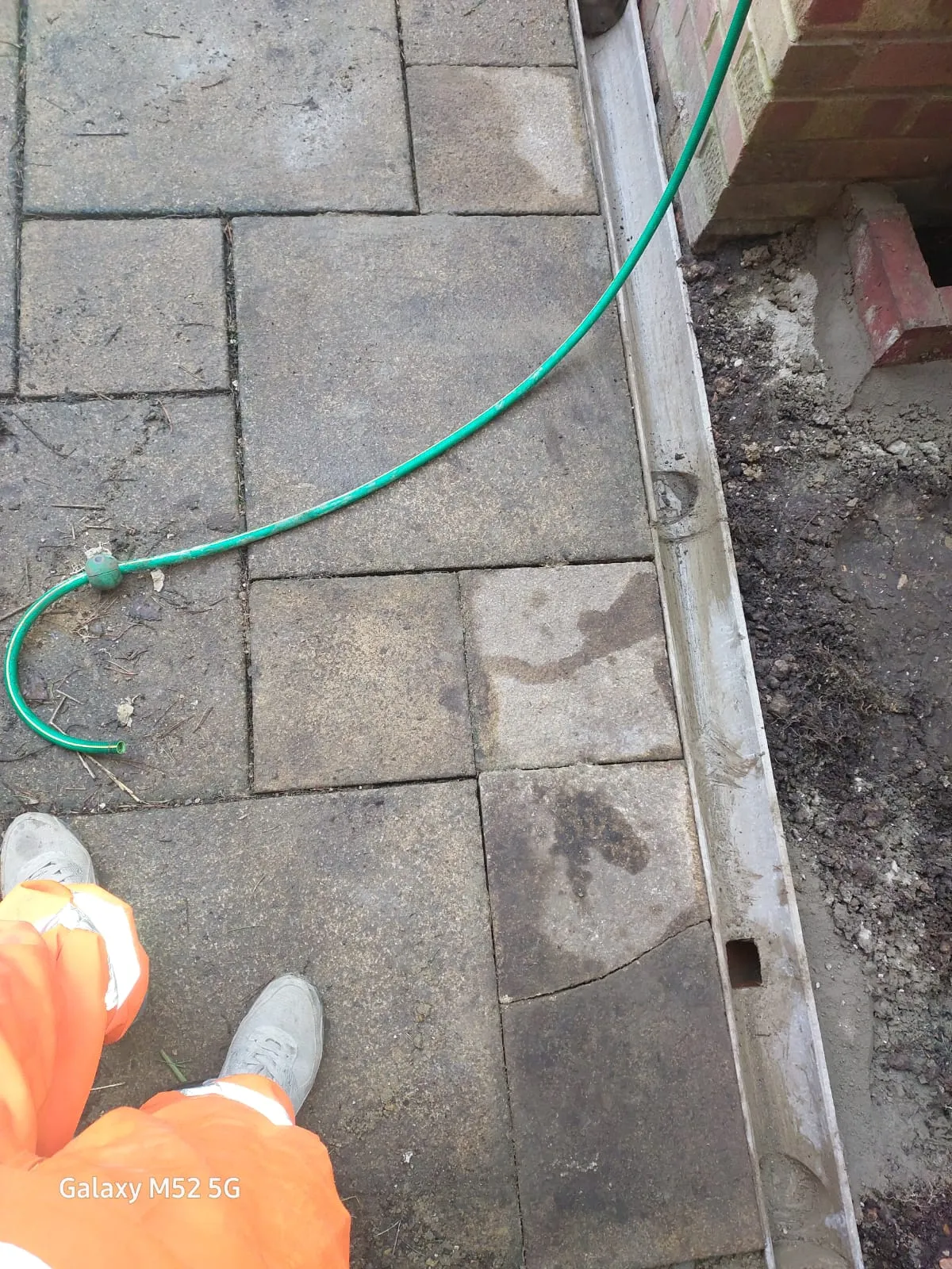 An ACO channel drain installation or repair in progress next to grey paving slabs and a brick wall, with a green hose and a worker's feet visible.