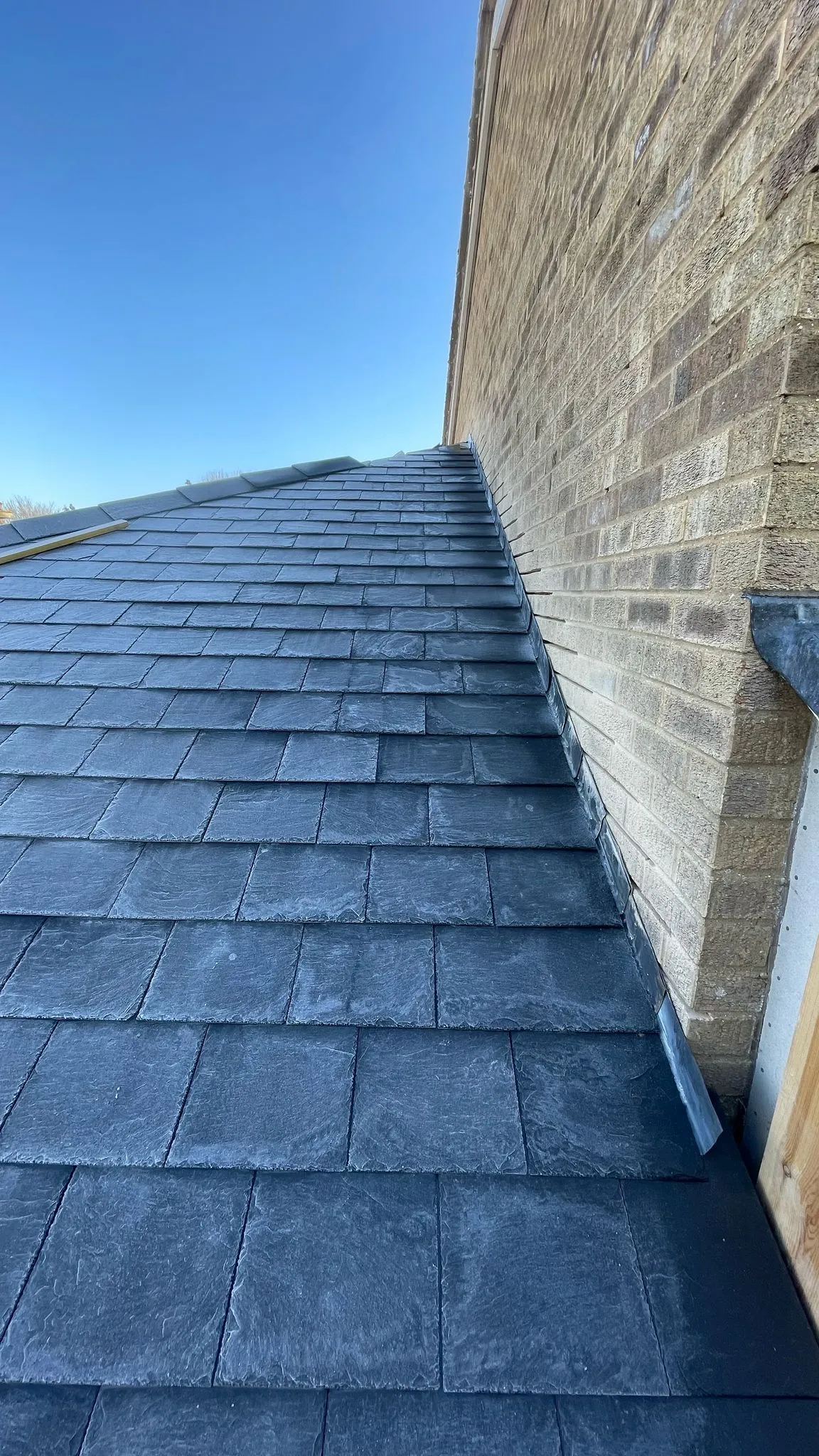 Newly installed dark slate roof tiles with lead flashing against a light brick wall under a blue sky.