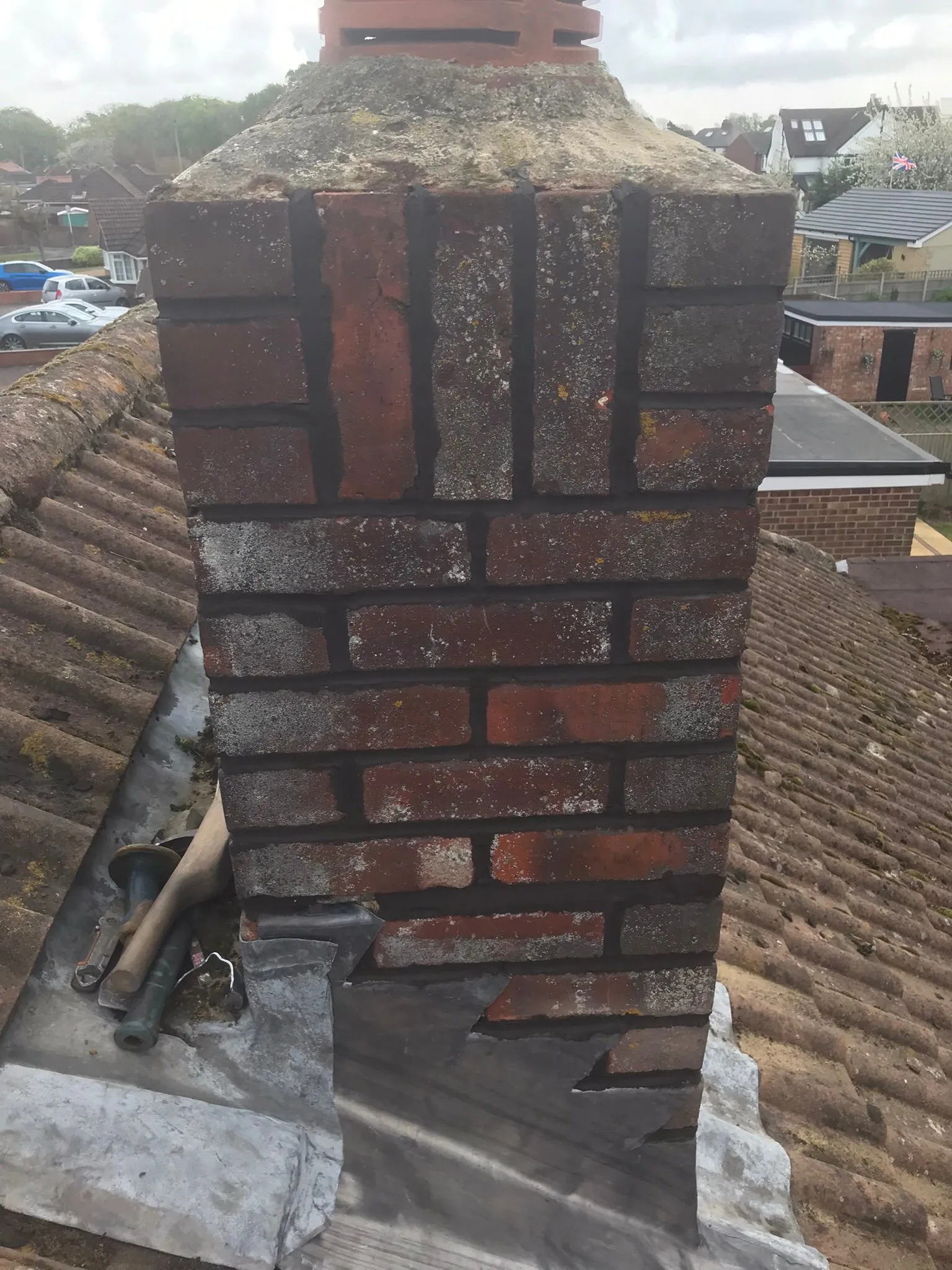 A brick chimney on a tiled roof with visible lead flashing, showing wear and moss, indicating a need for repair.