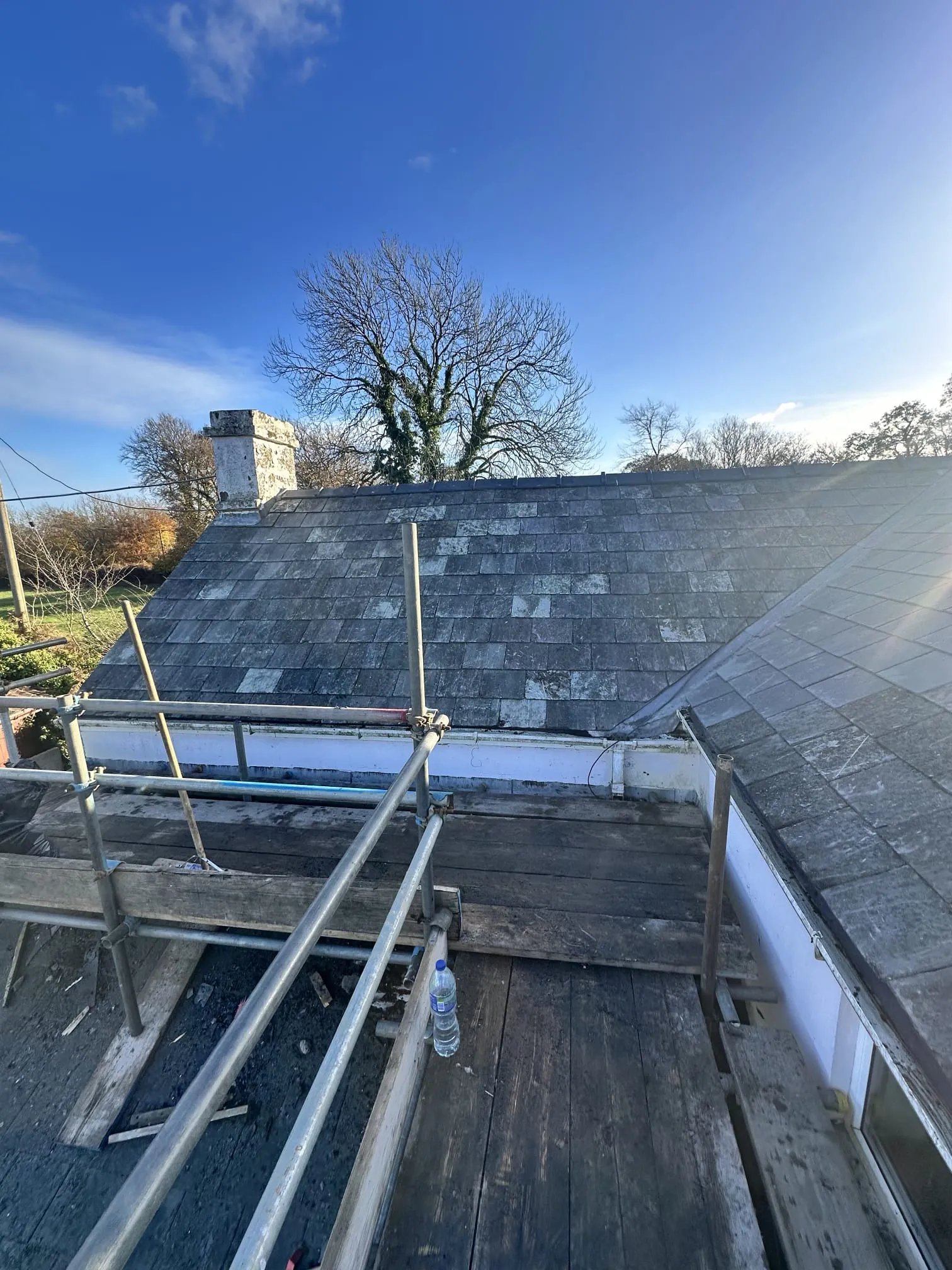 An old residential pitched roof with worn grey slate or tiles, a stone chimney, and scaffolding in the foreground under a bright blue sky, indicating ongoing roofing work.