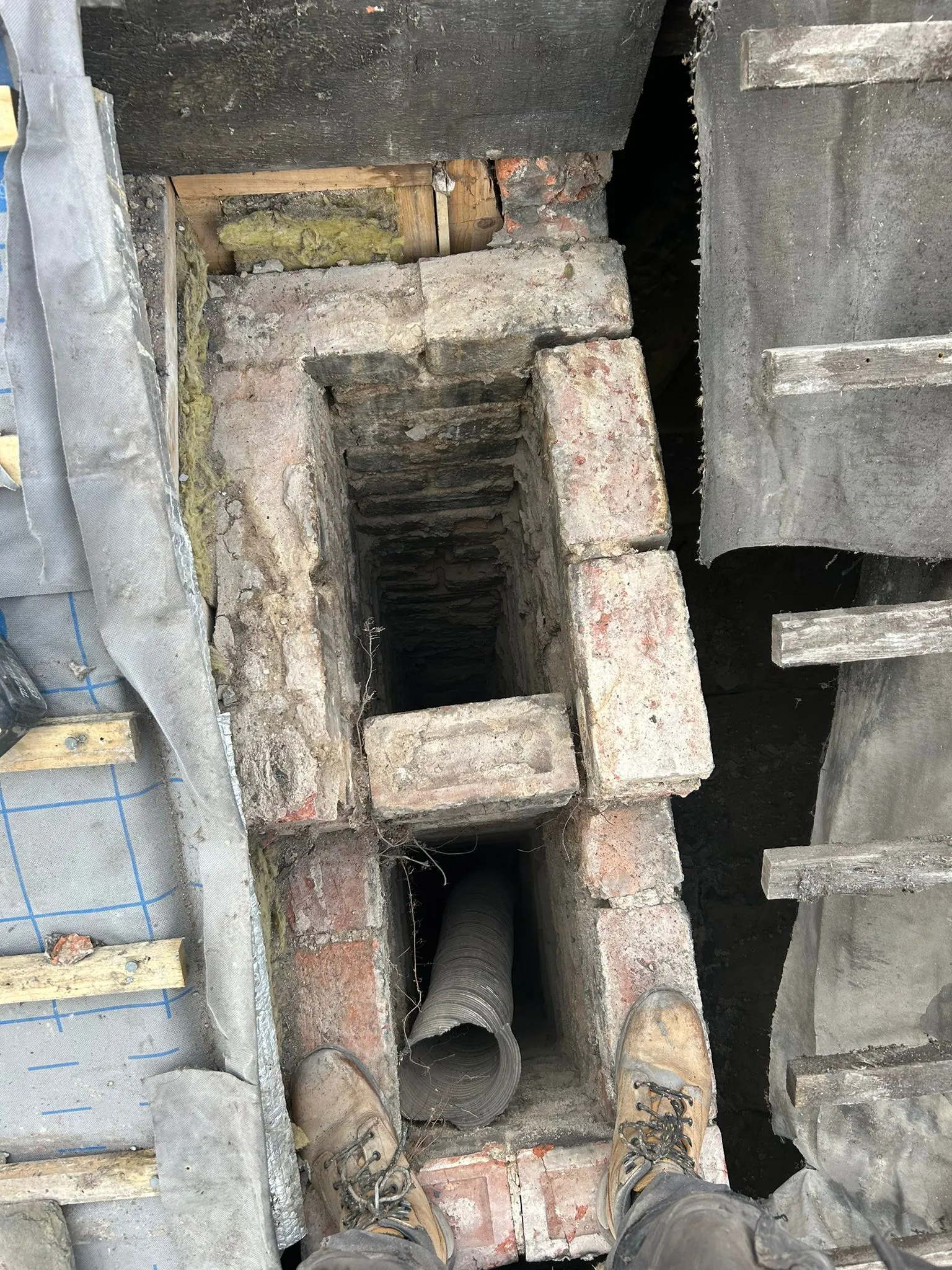 Looking down into an old brick chimney on a roof with visible flue liner and worker's boots.