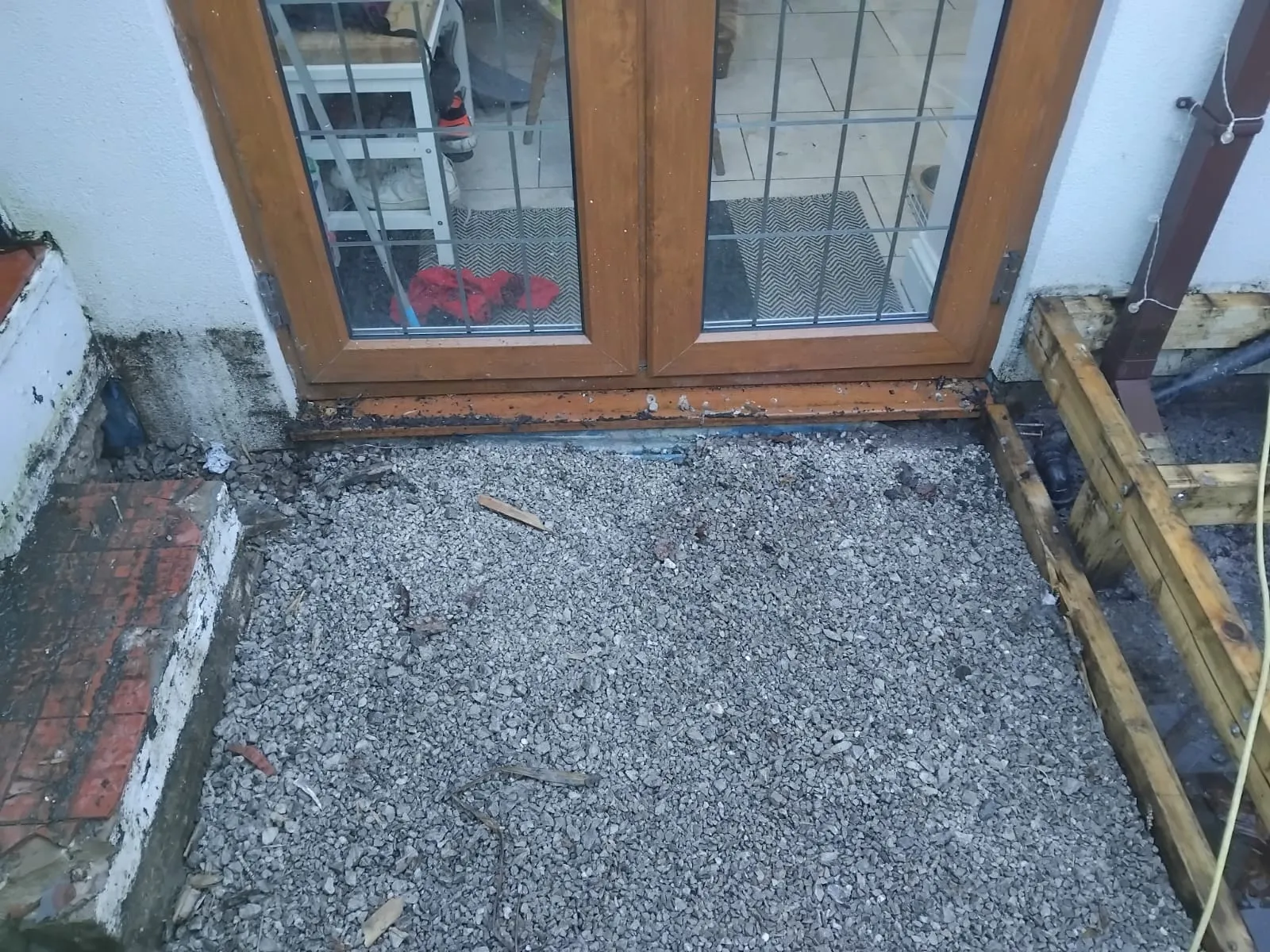 Residential property exterior showing a damp foundation, exposed gravel, and a downspout, suggesting a drainage issue needing repair or new installation.