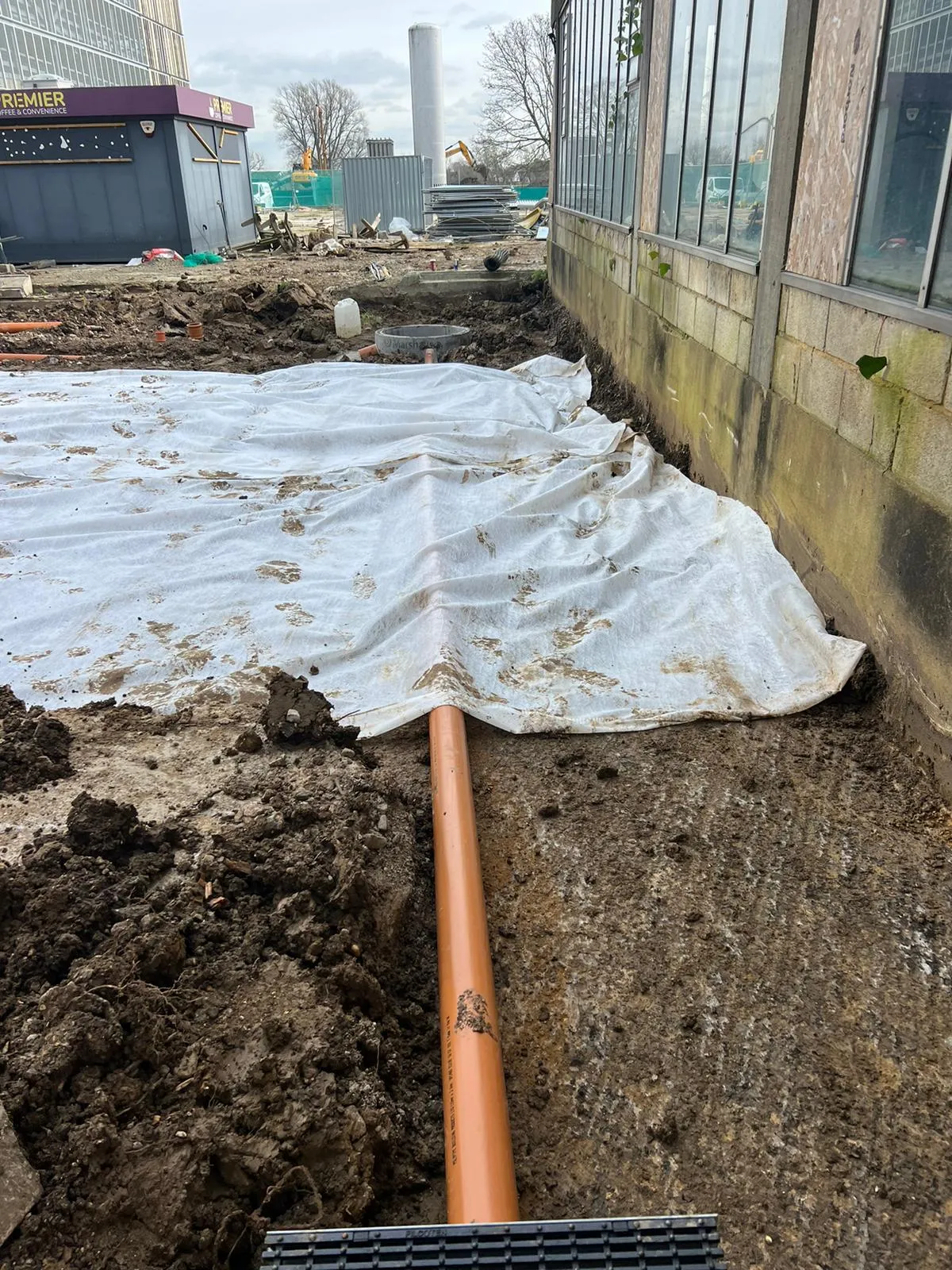 An orange plastic drainage pipe laid in an excavated trench next to a building, with white geotextile fabric partially covering the ground, indicating ongoing drainage installation or repair work.