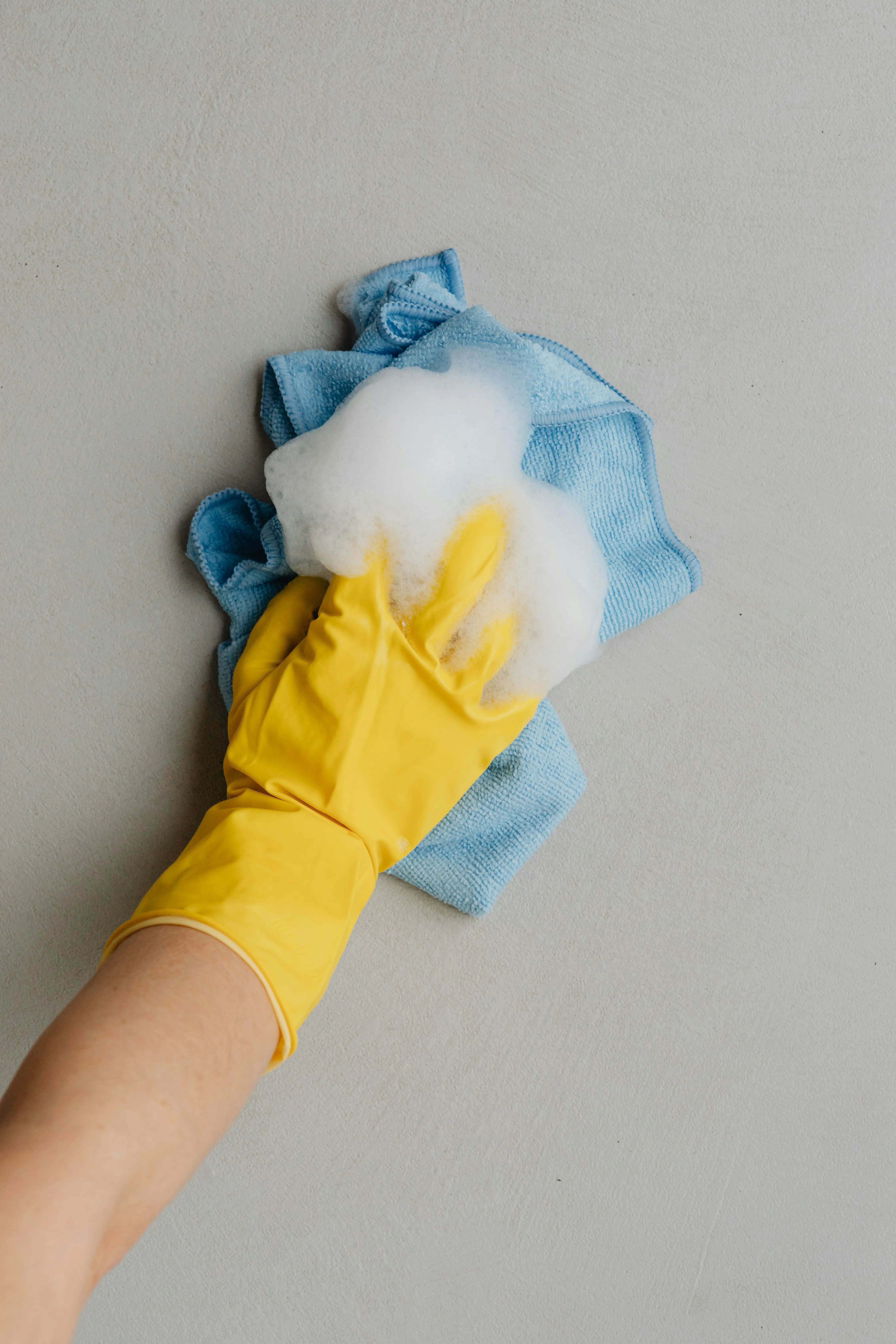 A hand wearing a yellow rubber glove holds a blue microfiber cloth covered in cleaning foam against a light grey background.