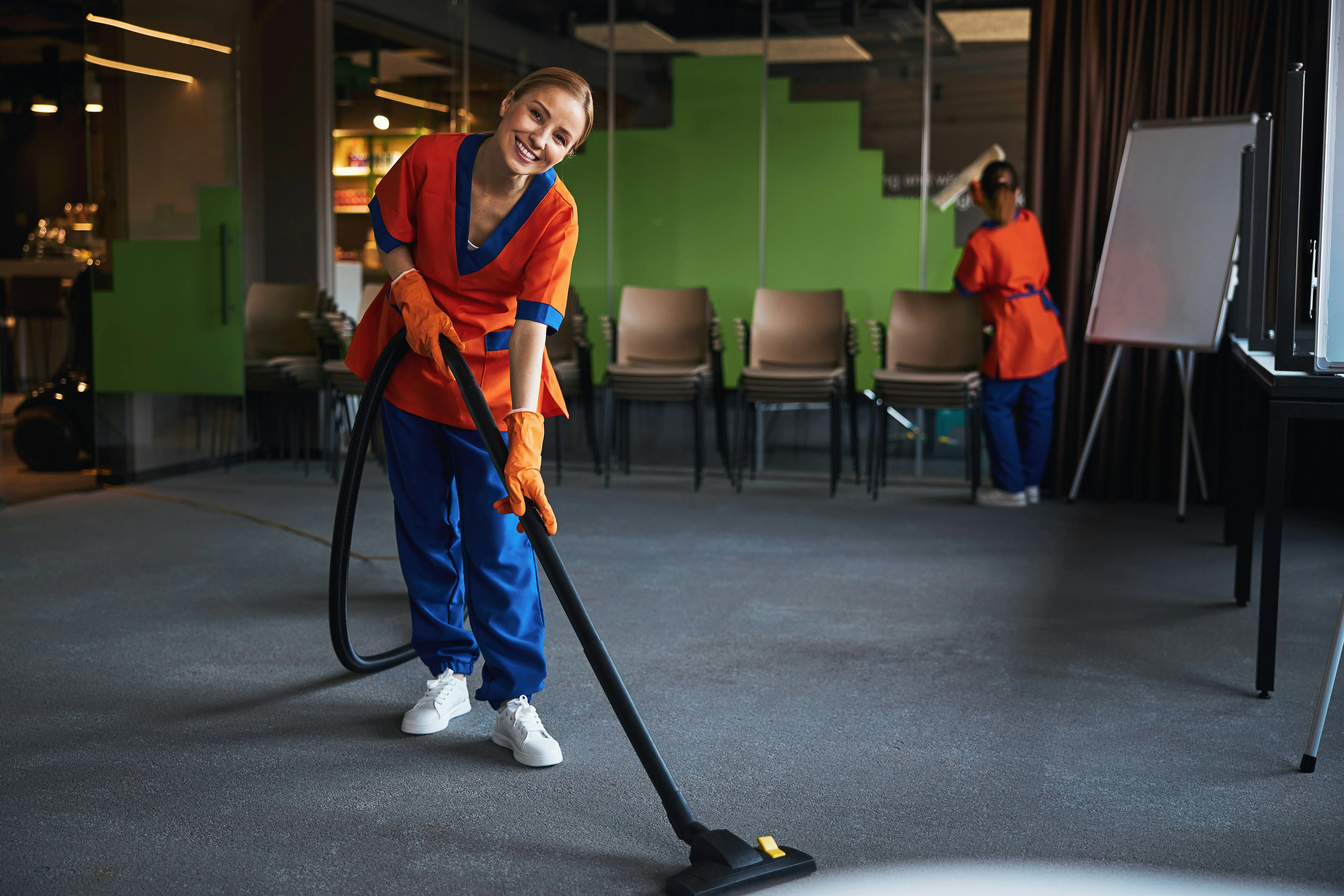 Two uniformed cleaners providing professional commercial office cleaning services, with one vacuuming a carpeted floor and another cleaning a window.