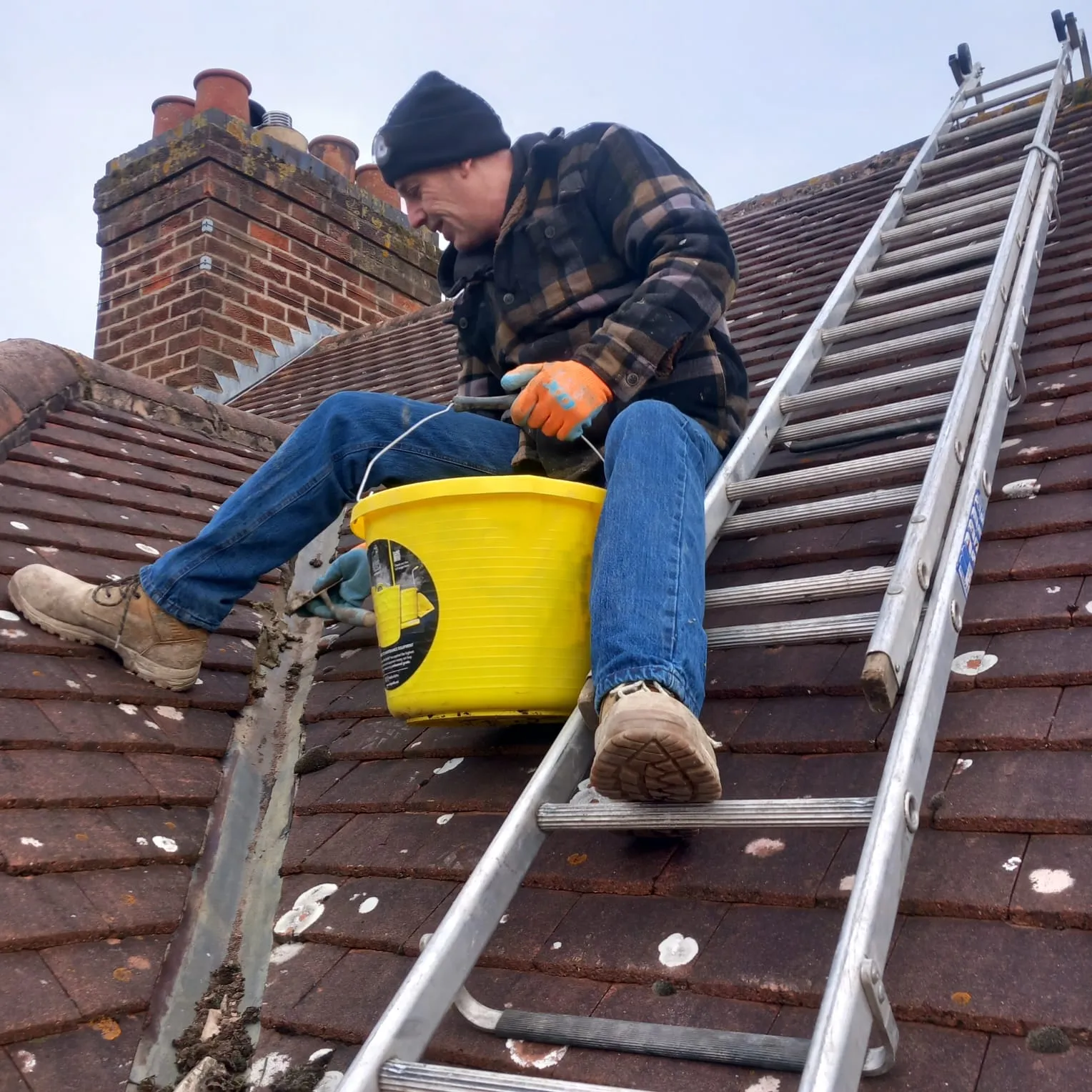 A professional roofer from 5 Star Roofing Leicester on a tiled roof, clearing a roof valley near a chimney with a yellow bucket.