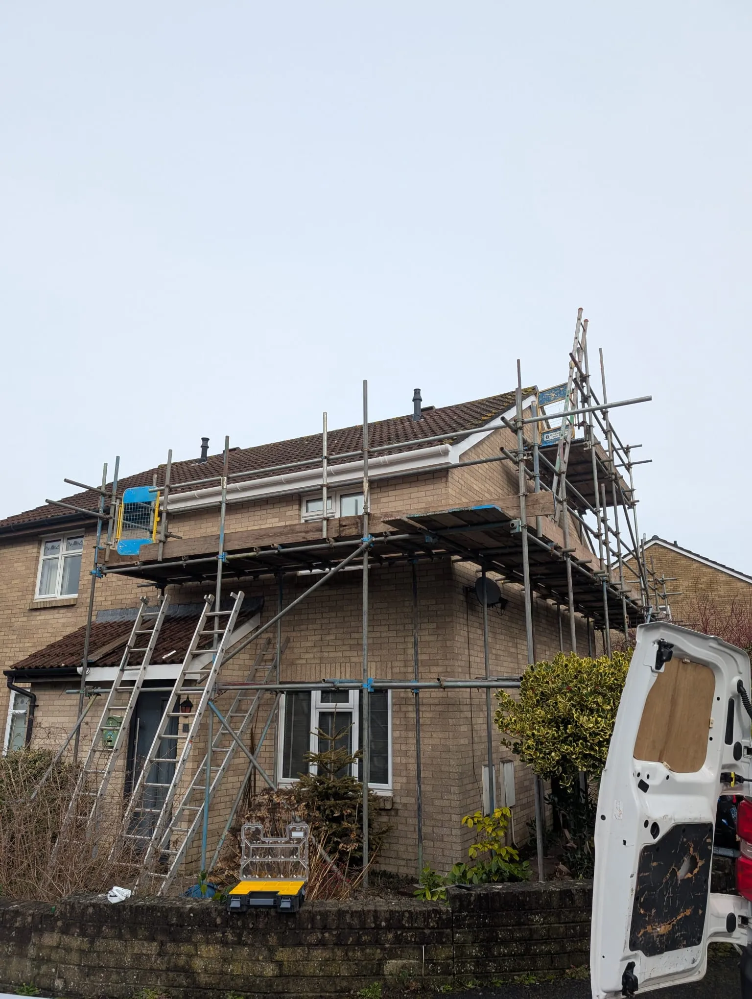 A brick residential house with full scaffolding for a re-roofing or roofline replacement project, showing new white gutters and a tiled roof.