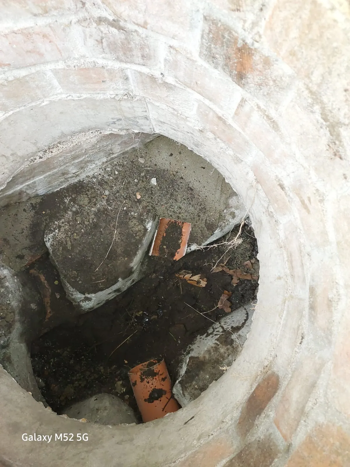 An overhead view inside a drainage manhole showing accumulated debris, dirt, and sections of broken orange pipe.