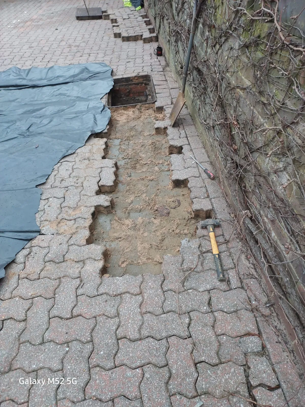Paving blocks removed to reveal an open drainage access point and soil beneath, with tools nearby, signifying ongoing drain repair or installation.