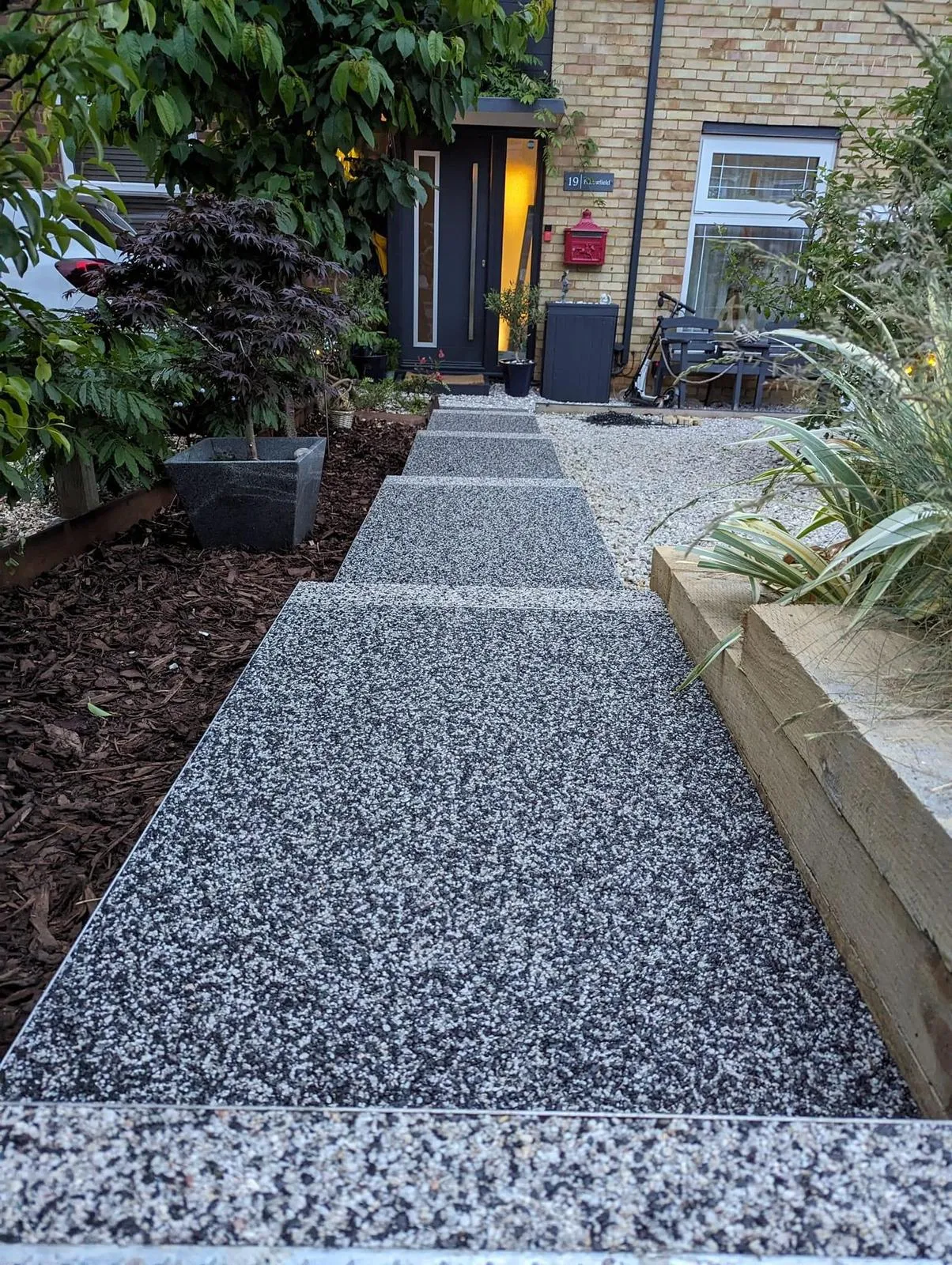 A contemporary resin-bound pathway with steps, bordered by garden landscaping featuring bark and a gravel bed, leading to a residential entrance.