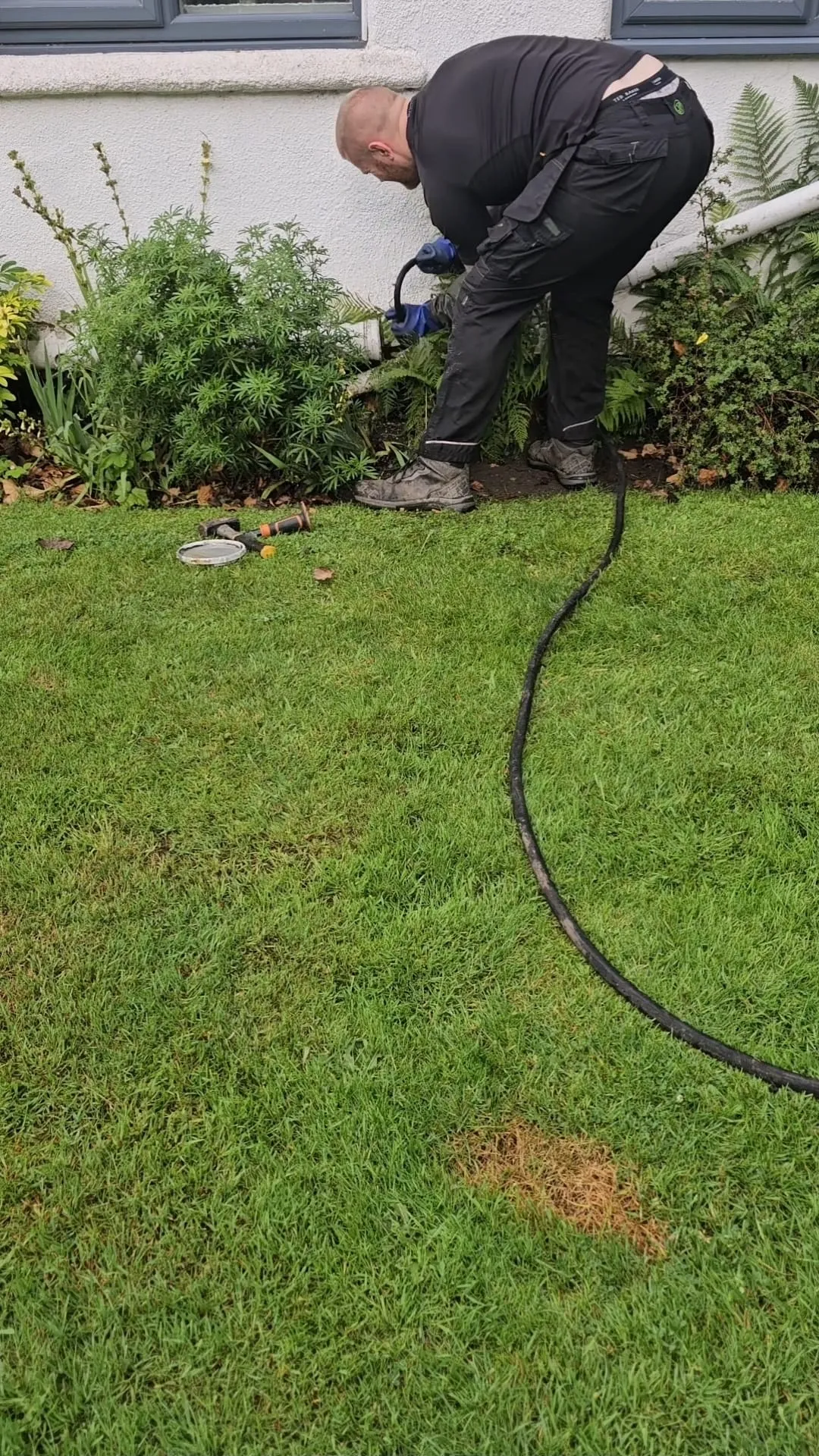A drainage technician in work clothes clearing an outdoor drain next to a house.