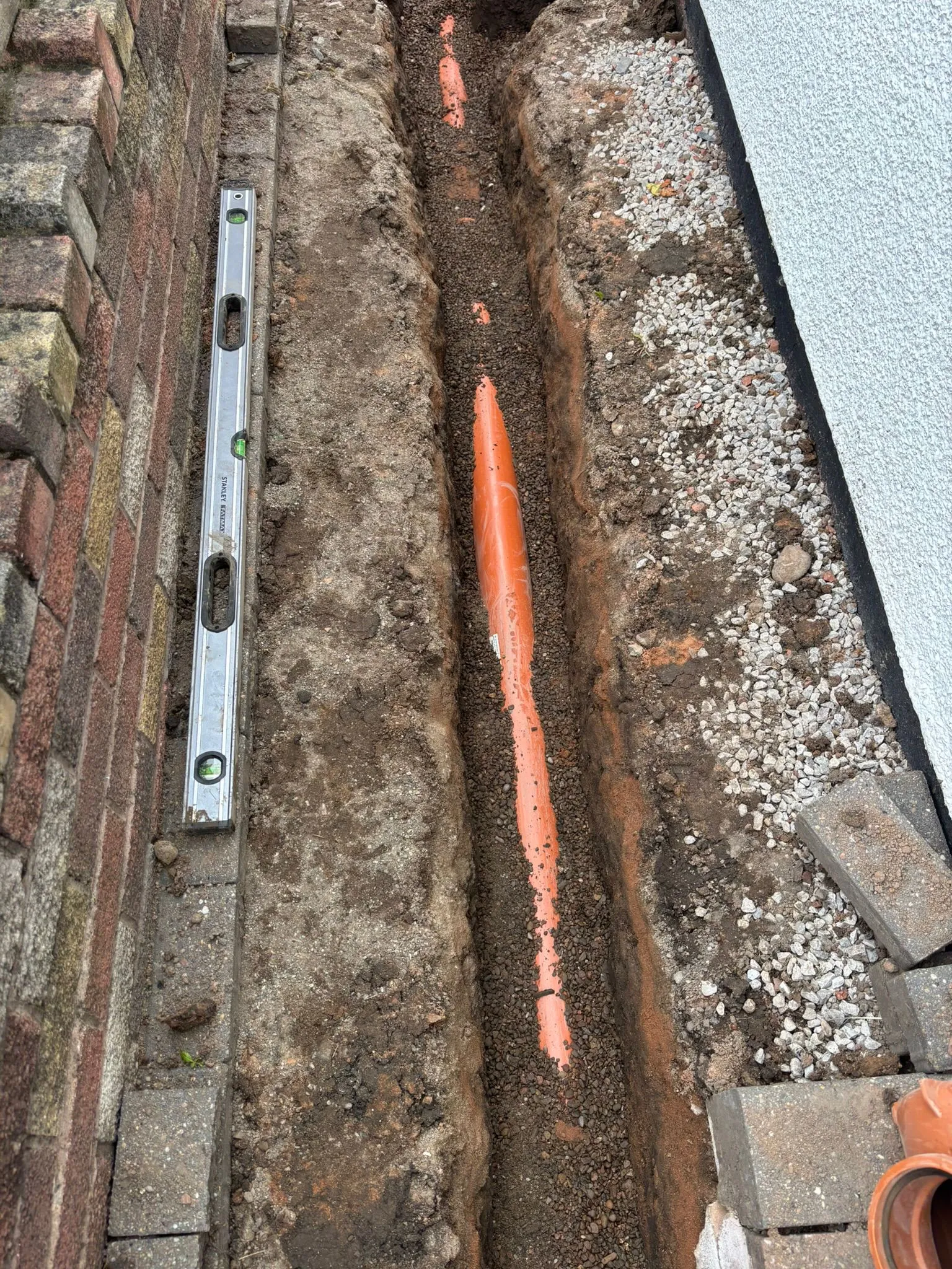 Drainage pipe installation in an excavated trench with a spirit level, brick wall, and building.