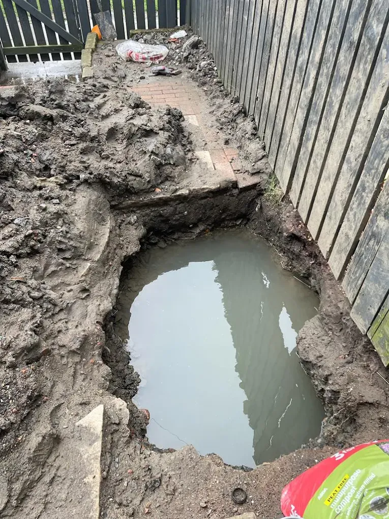 Photo showing a large hole dug in the ground filled with water, with muddy earth around it, alongside a fence and paving.