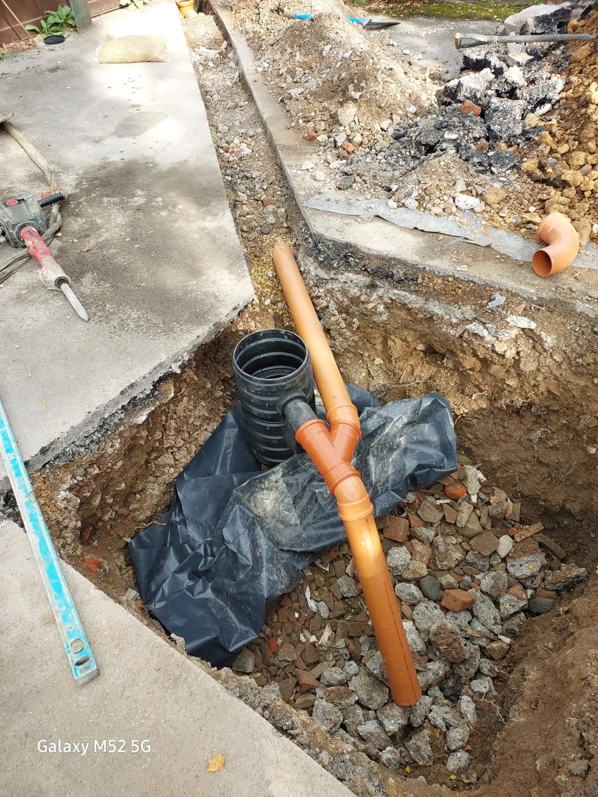 An open trench showing newly laid orange plastic drainage pipes and a black corrugated inspection chamber, with a jackhammer and soil piles nearby, indicating ongoing drain installation or repair work.
