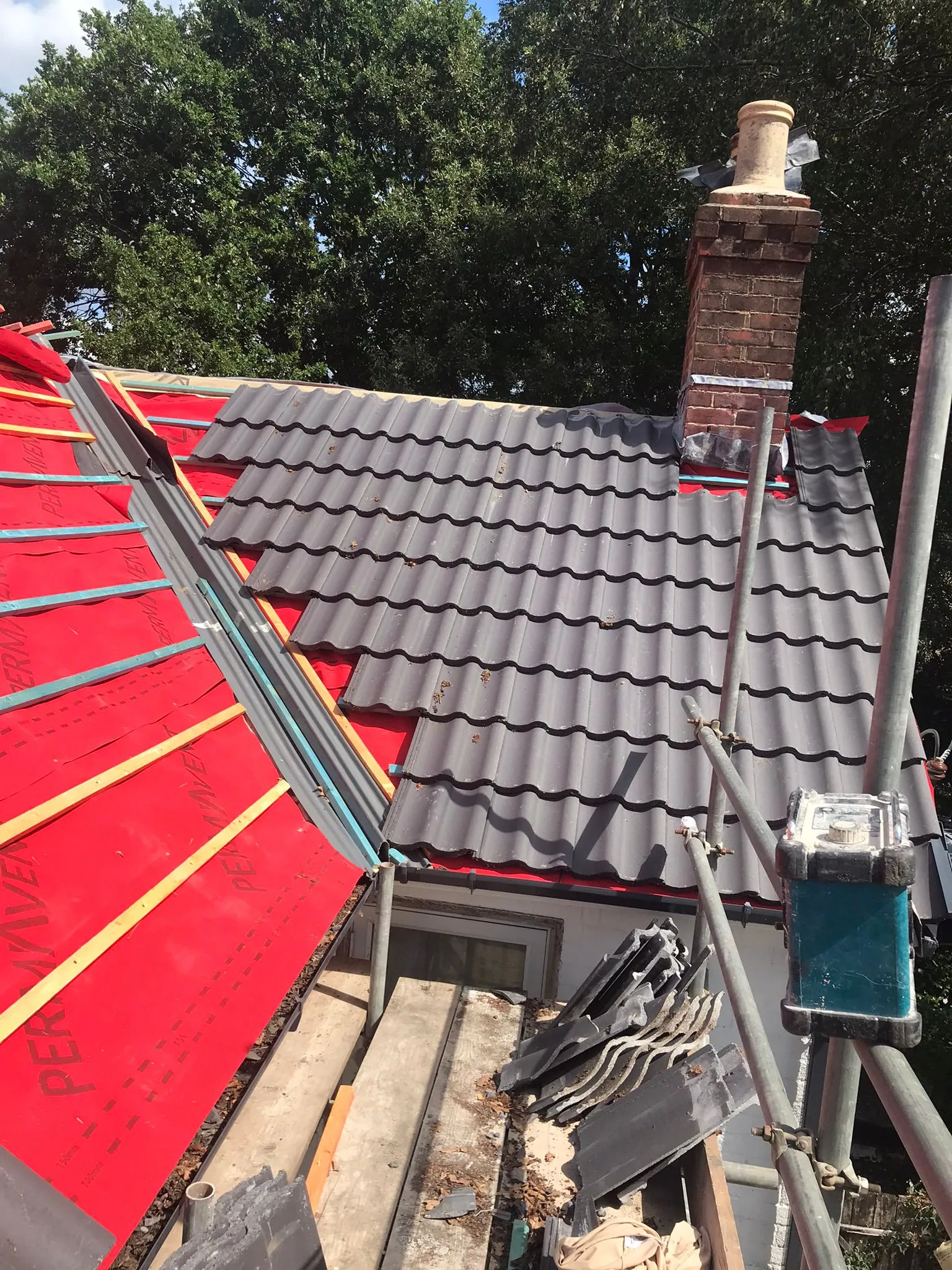 Roof replacement or installation in progress on a residential property, showing new grey tiles, red roofing felt, and a brick chimney.