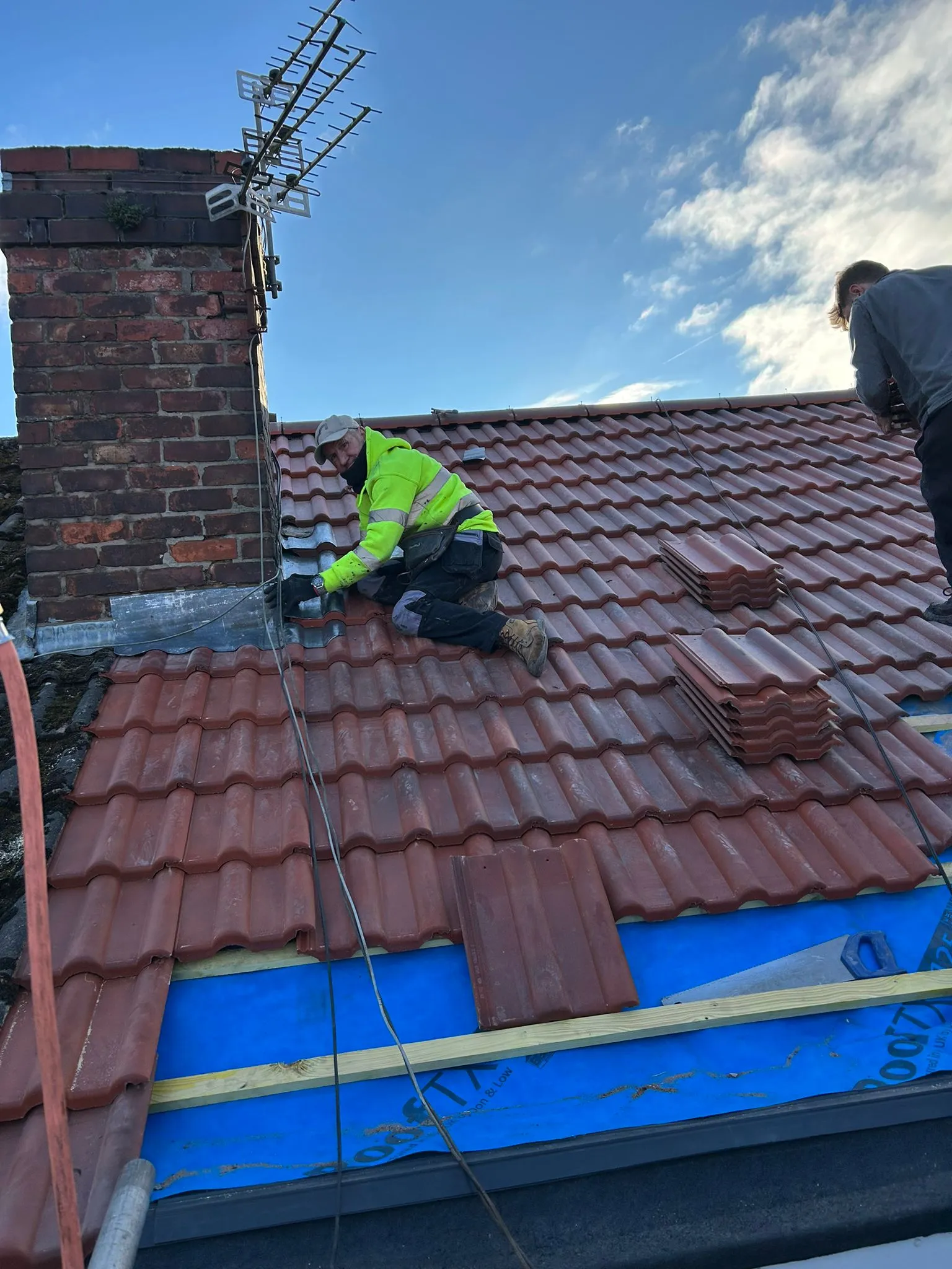 Roofers installing new red tiles and lead flashing on a pitched roof with a chimney