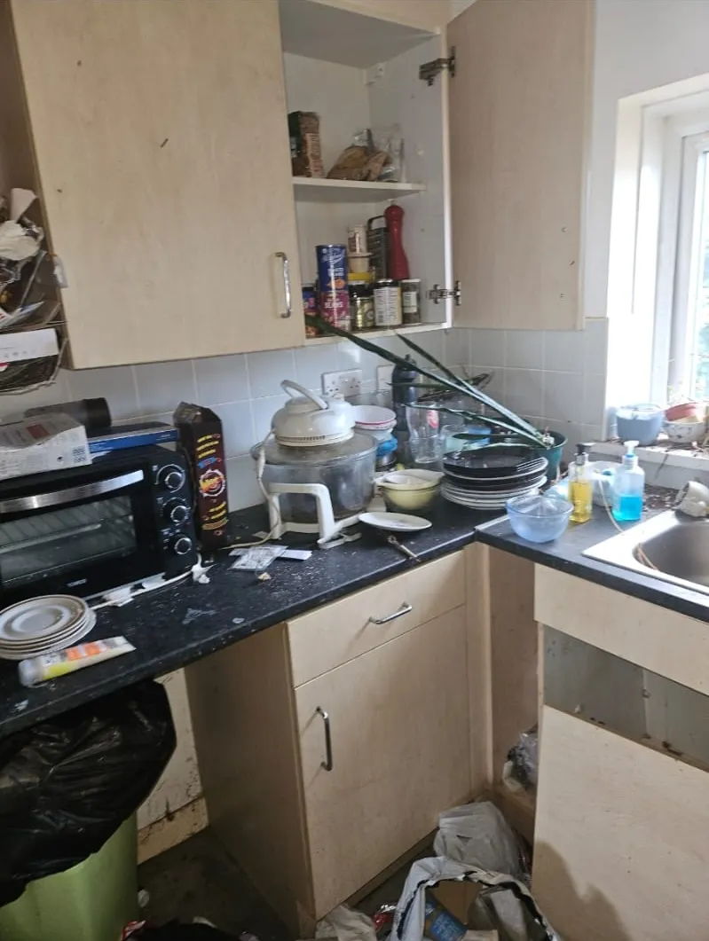 A heavily cluttered and untidy kitchen with dirty dishes, open cabinets, and rubbish on the floor, ready for a house clearance service.