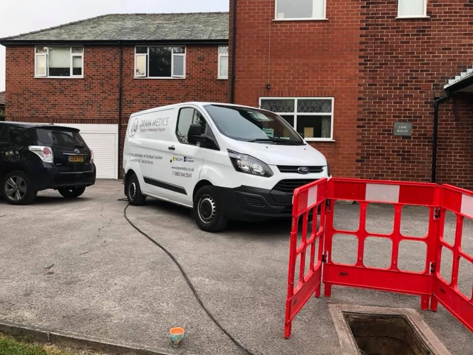 Drain Medics van with a hose near an open manhole and safety barriers on a residential driveway.
