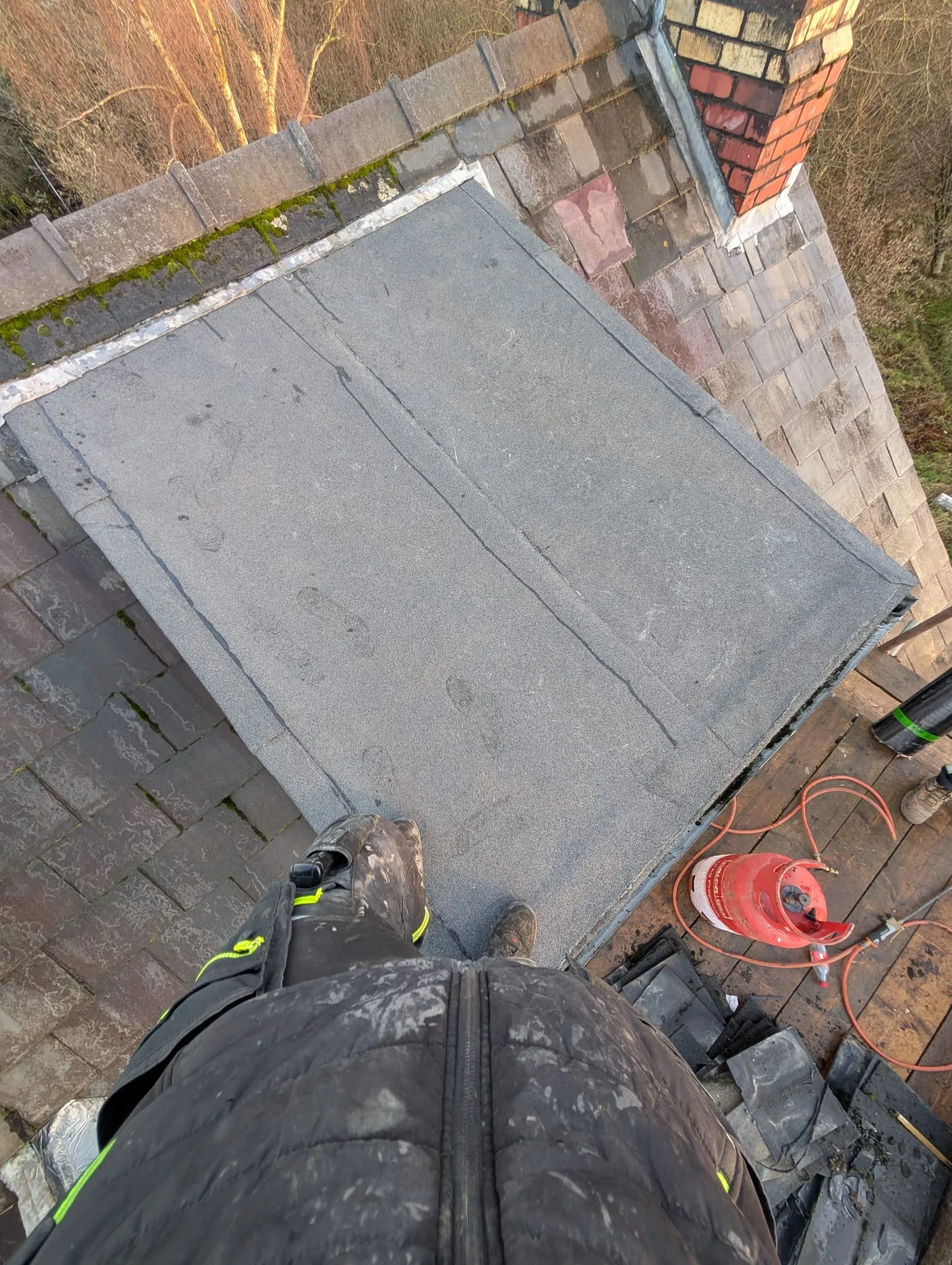 Roofer's perspective of a flat felt roof installation or repair, next to an old slate roof and a brick chimney, with a gas bottle and tools on a wooden platform.