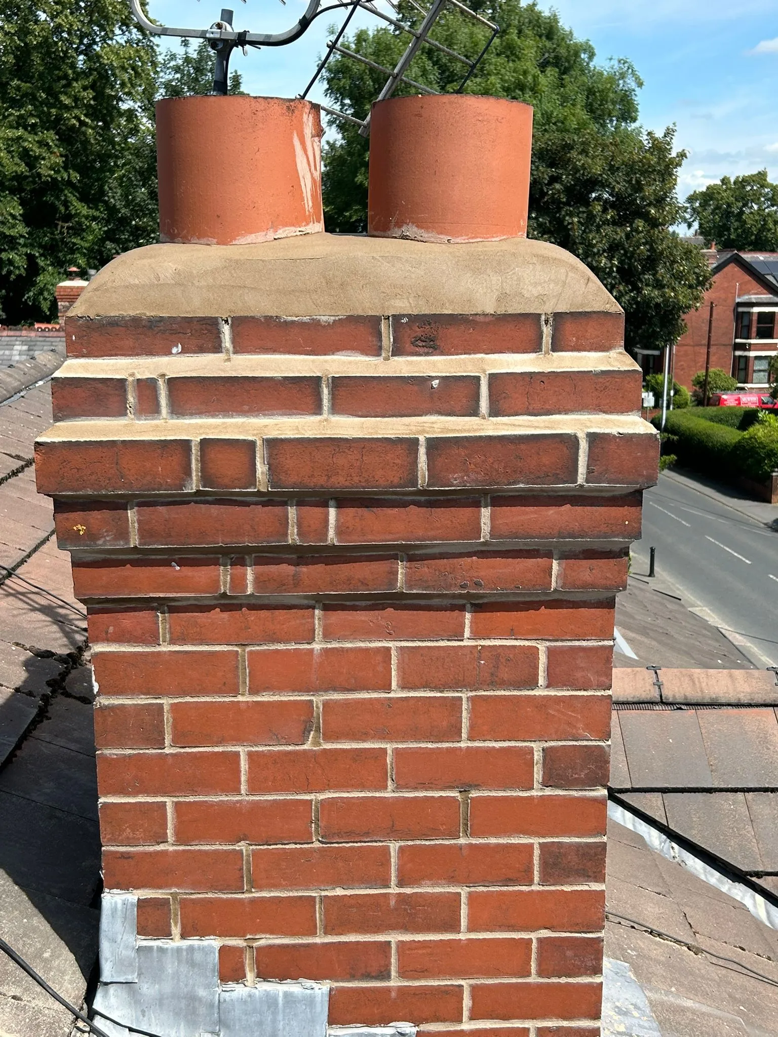 A close-up of a well-maintained brick chimney with repointed mortar, lead flashing, and two terracotta pots on a tiled roof.