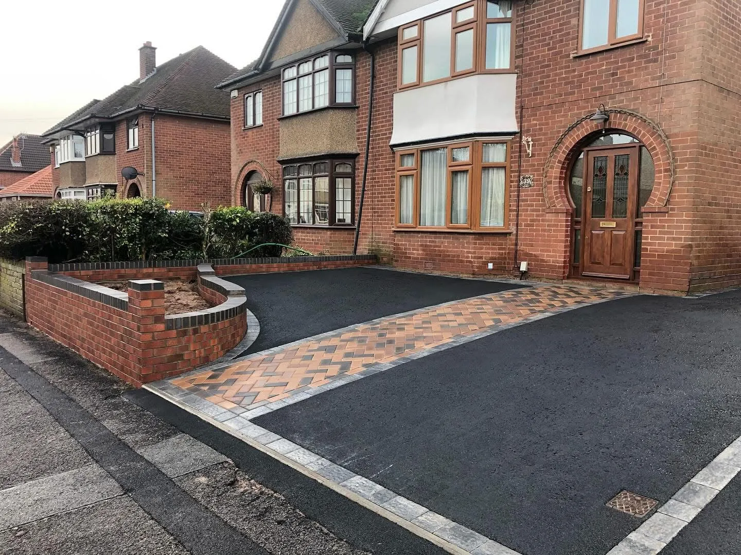 Newly laid tarmac driveway with a decorative block paving pathway and a curved brick retaining wall in front of a red brick house.