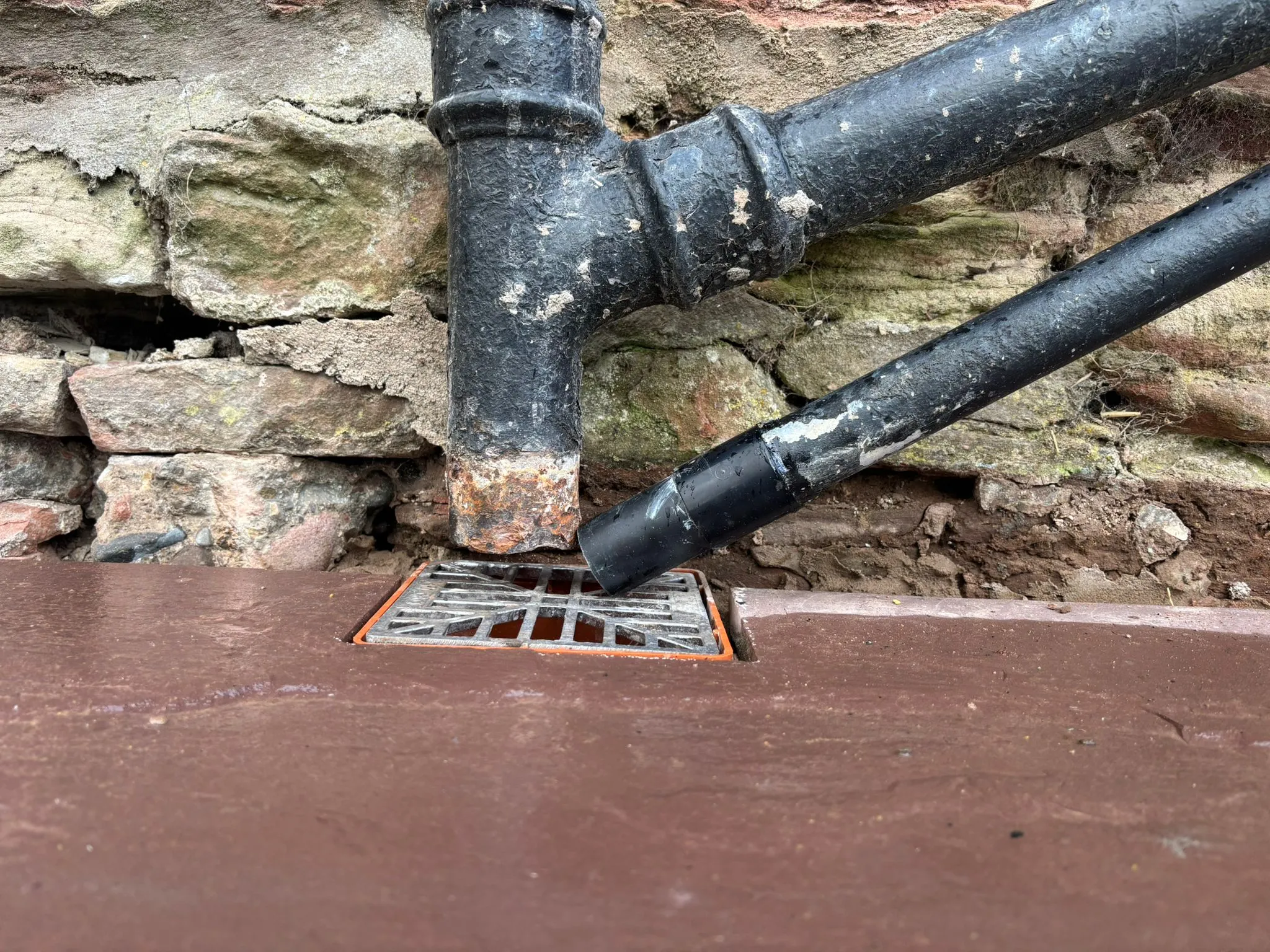 An outdoor drain with two black drainage pipes leading into it, set against a rustic stone wall.