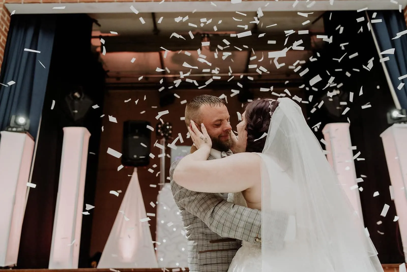 Bride and groom embracing under a shower of white confetti during their wedding reception, with event lighting in the background.
