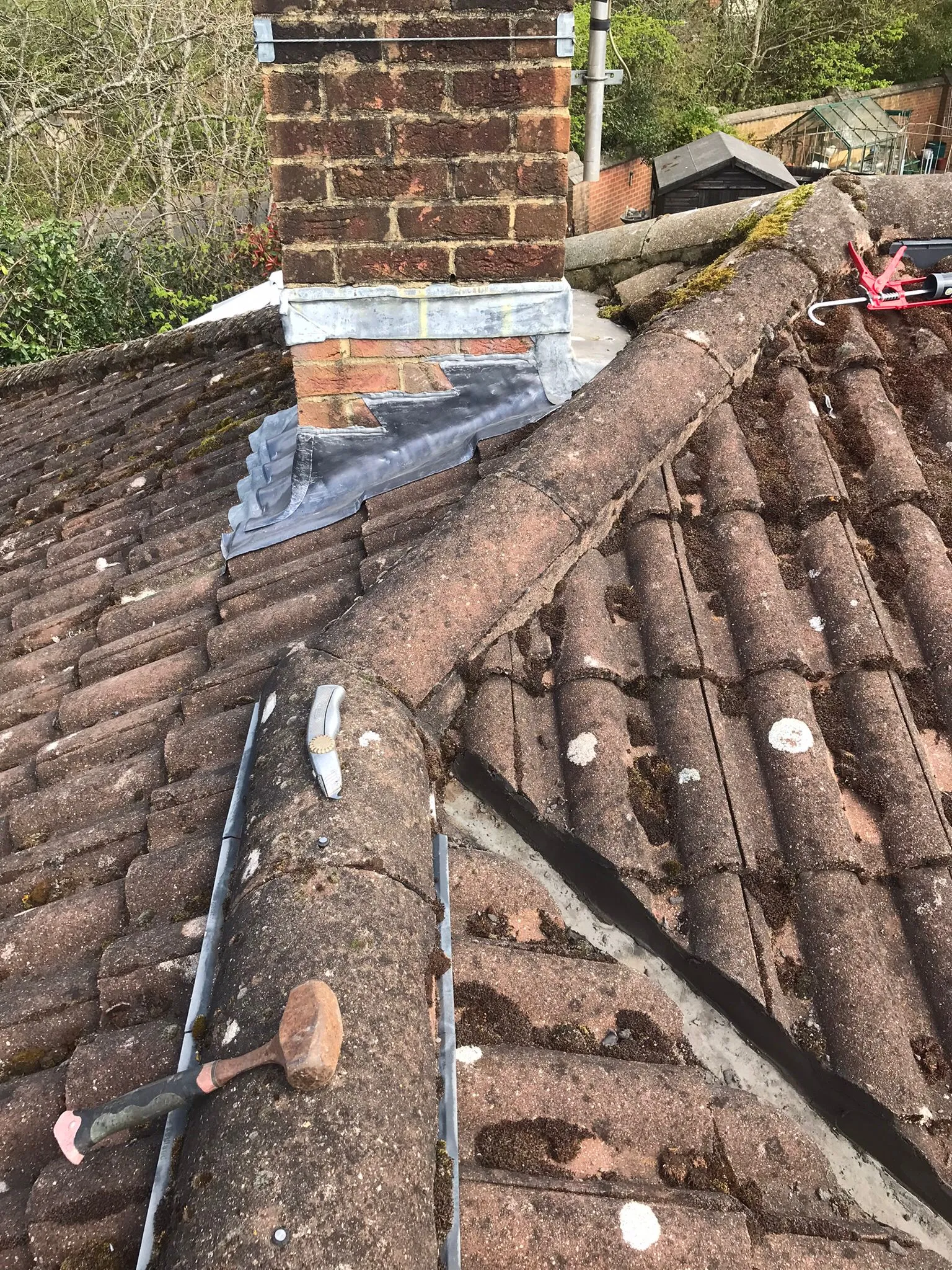 Tiled roof with moss, a brick chimney stack with lead flashing, and roofing tools like a hammer and caulking gun.