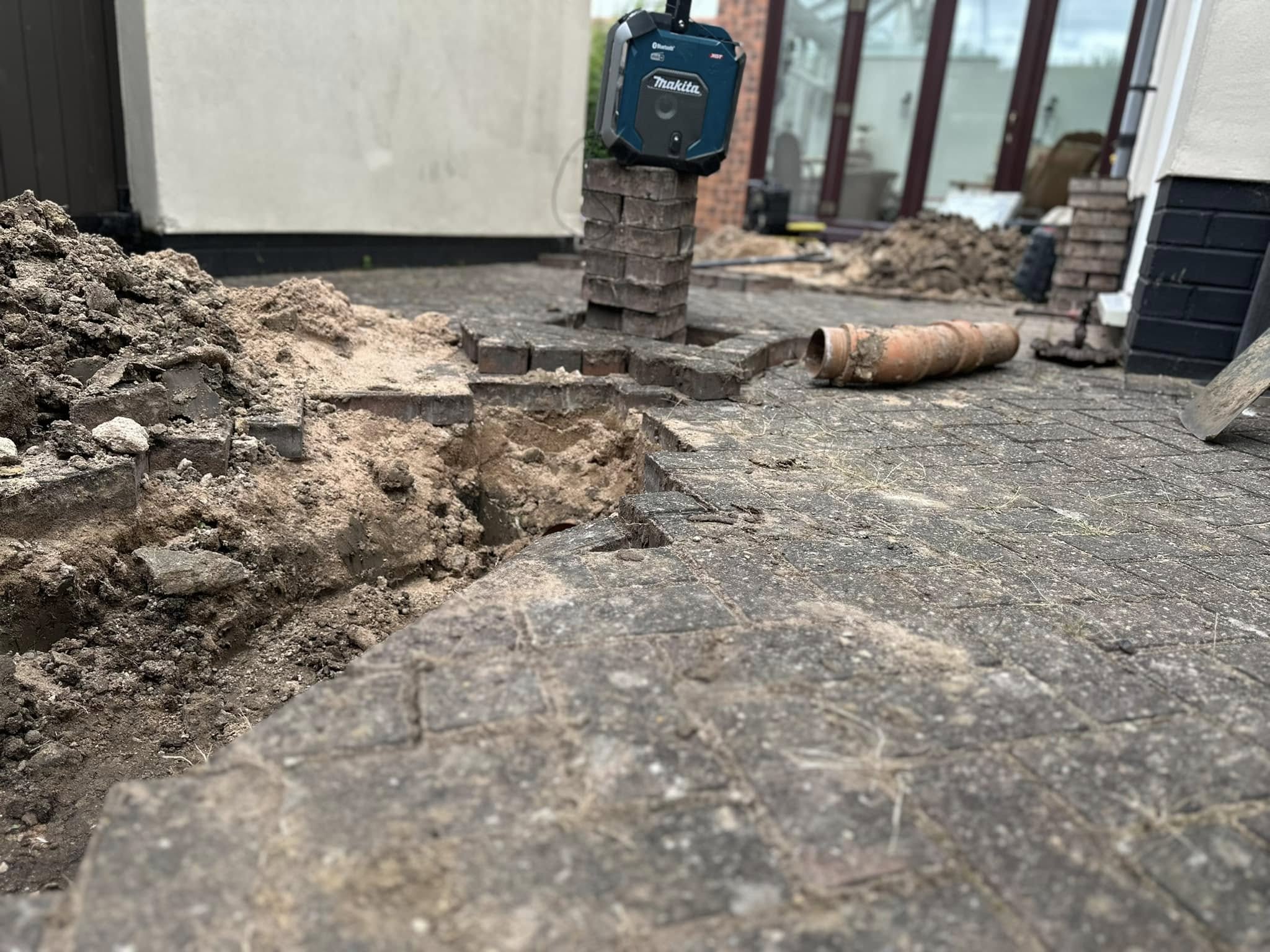 Drainage repair work in progress, showing an excavated hole in grey block paving with soil and rubble piles, a removed orange drain pipe, and a Makita laser level on a brick stack.
